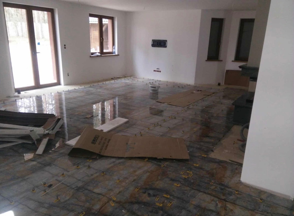 Układanie płytek podłogowych w jasnym wnętrzu. Widoczne narzędzia glazurnicze i elementy kartonowe zabezpieczające podłoże. Płytki z efektem marmuru.