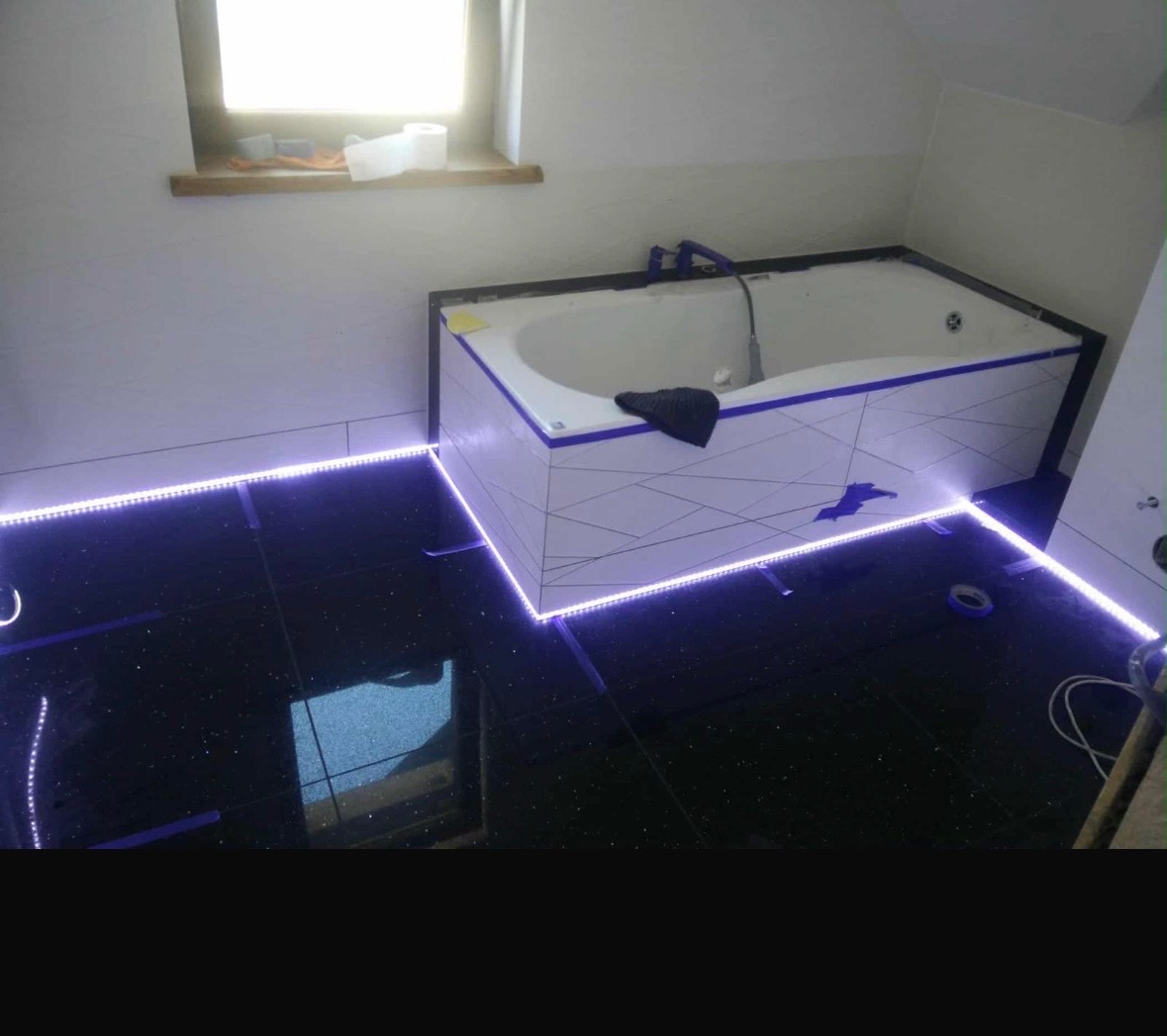 Wykończona łazienka z czarną, błyszczącą podłogą i podświetloną wanną. Widoczne taśmy LED i fragment okna z akcesoriami łazienkowymi. Ukończony remont łazienki.