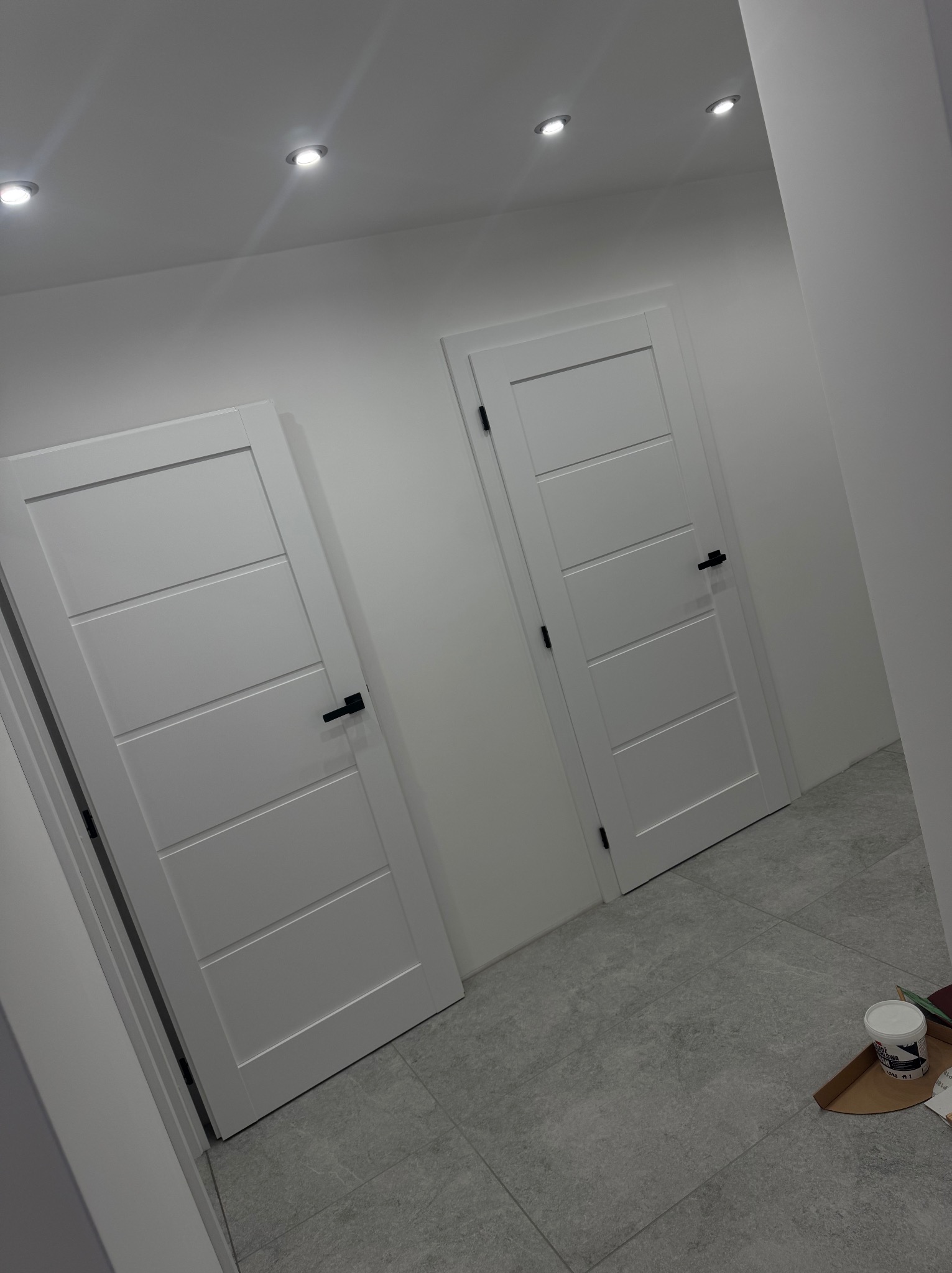 Dwa białe, nowoczesne drzwi z czarnymi klamkami w jasnym korytarzu z szarą podłogą. Widoczne punktowe oświetlenie sufitowe i akcesoria malarskie.