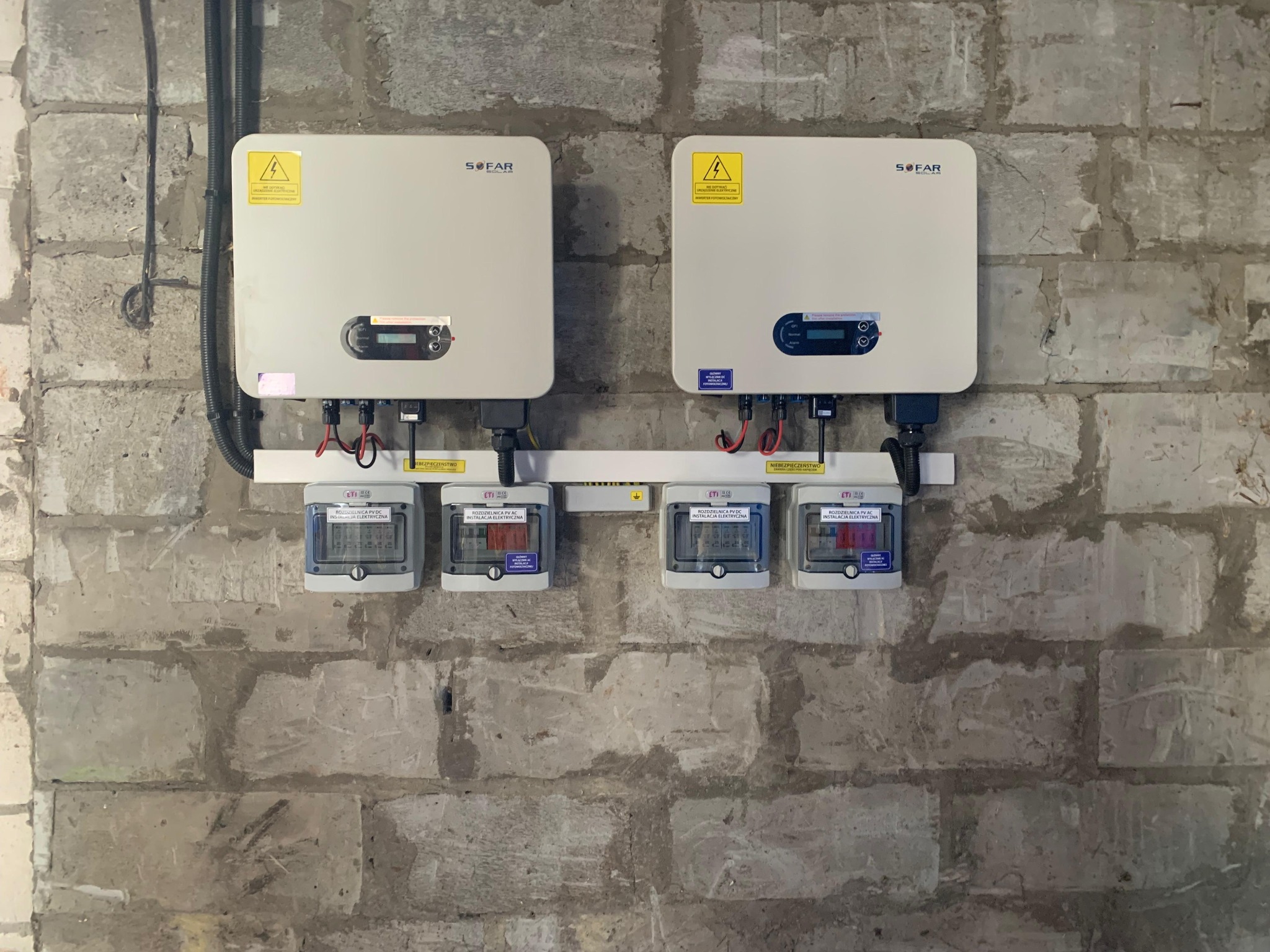Dwa inwertery SOFAR i cztery rozdzielnice PV AC/DC zamontowane na szarej, surowej ścianie z cegieł. Widoczne przewody i etykiety ostrzegawcze.