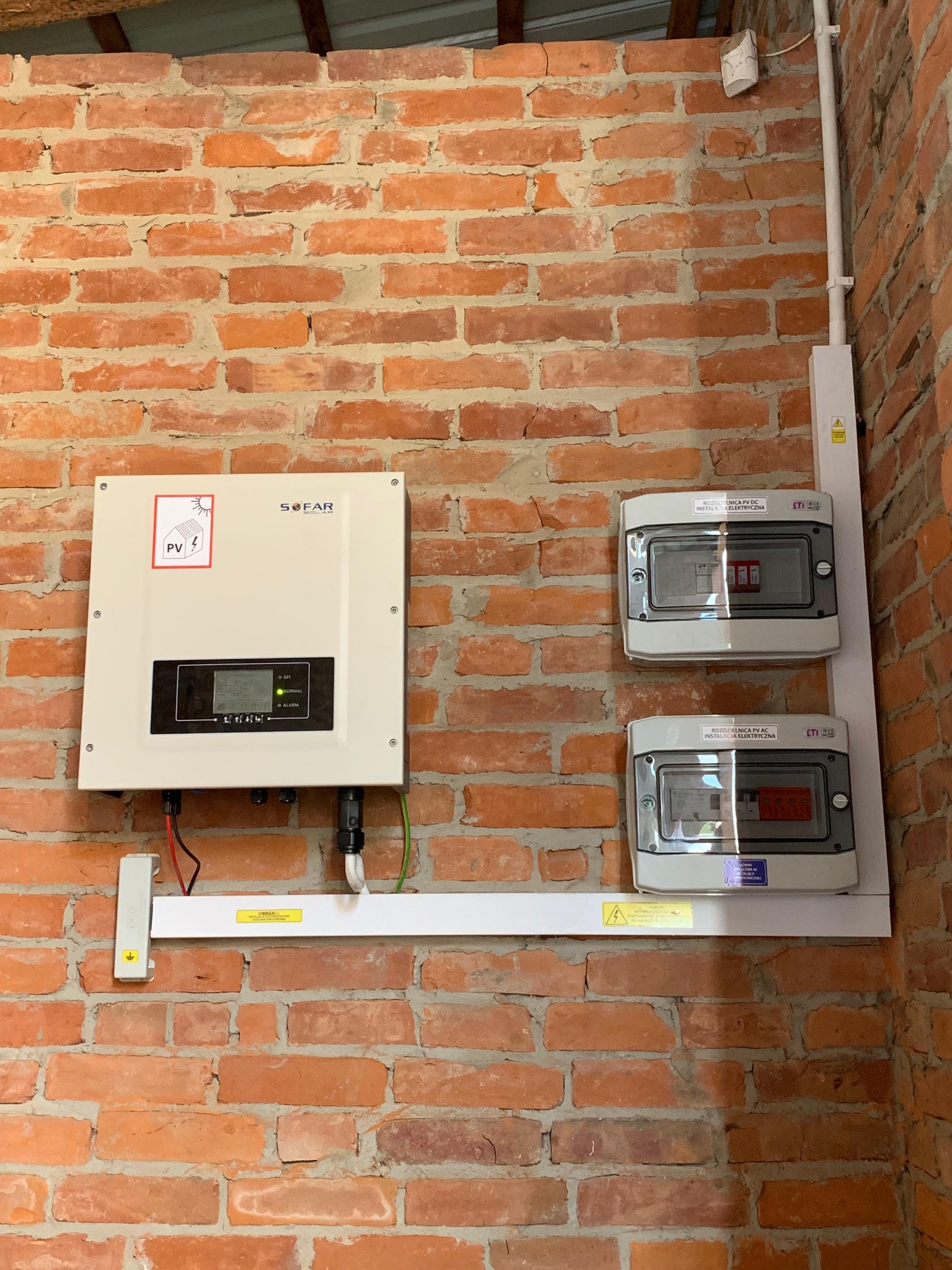 Instalacja fotowoltaiczna wewnątrz budynku z ceglaną ścianą. Widoczny inwerter Sofar i rozdzielnice PV AC/DC. Białe elementy instalacji kontrastują z czerwoną cegłą.