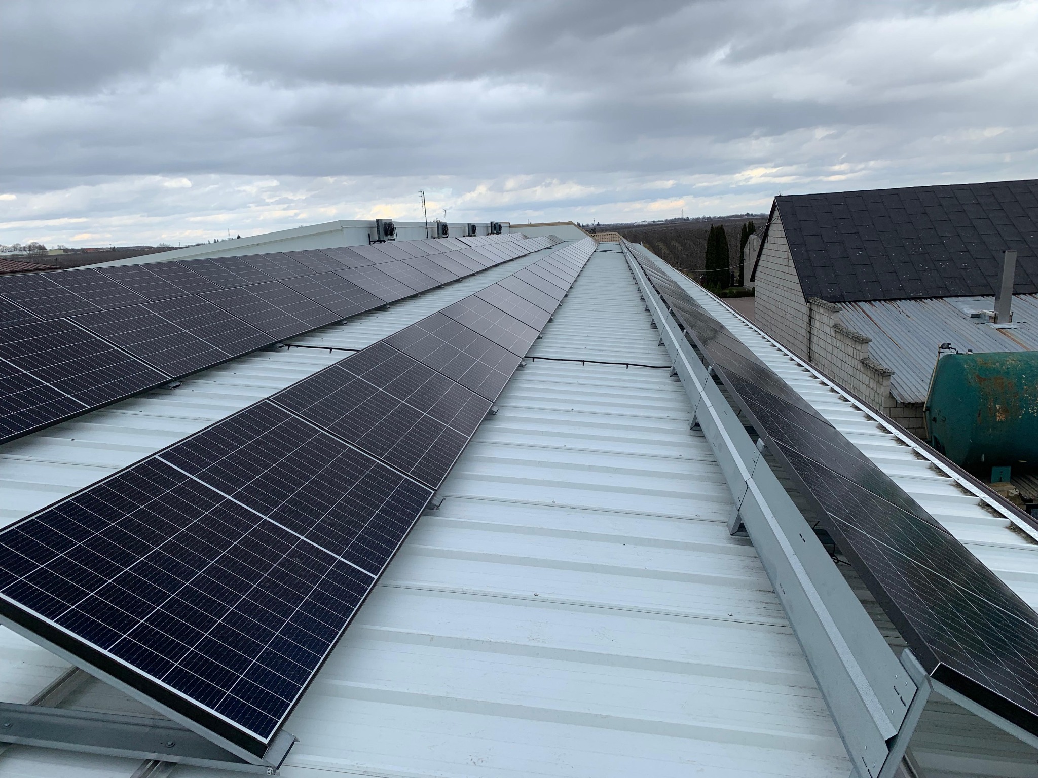 Rząd paneli słonecznych na blaszanym dachu budynku gospodarczego w pochmurny dzień. Widok z góry, panele zamontowane na metalowych konstrukcjach.