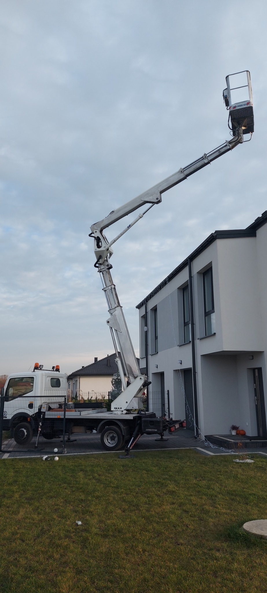 Podnośnik koszowy na tle nowoczesnego domu z dekoracjami świątecznymi. Widoczny kosz na wysięgniku i fragment trawnika z ozdobami.