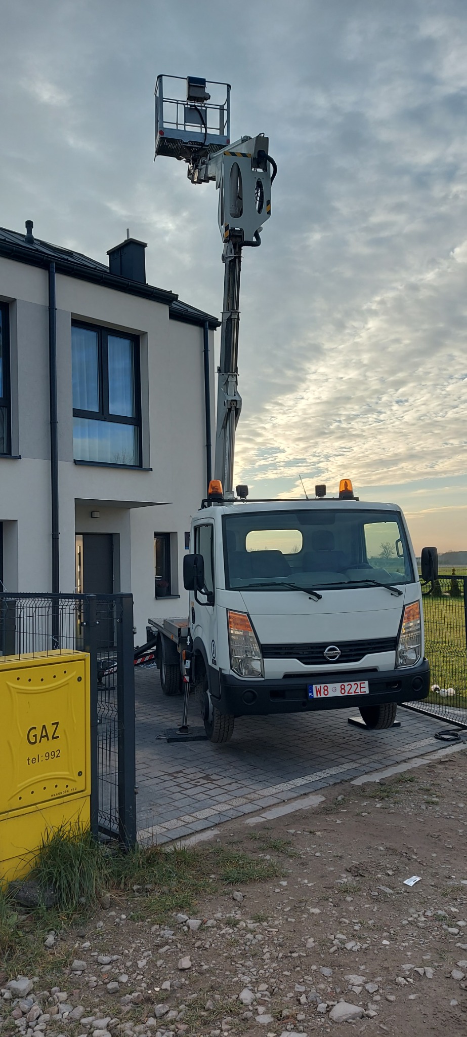 Podnośnik koszowy na chodniku przed nowoczesnym domem z szarymi oknami. Żółta skrzynka gazowa przy ogrodzeniu. Pochmurne niebo w tle.