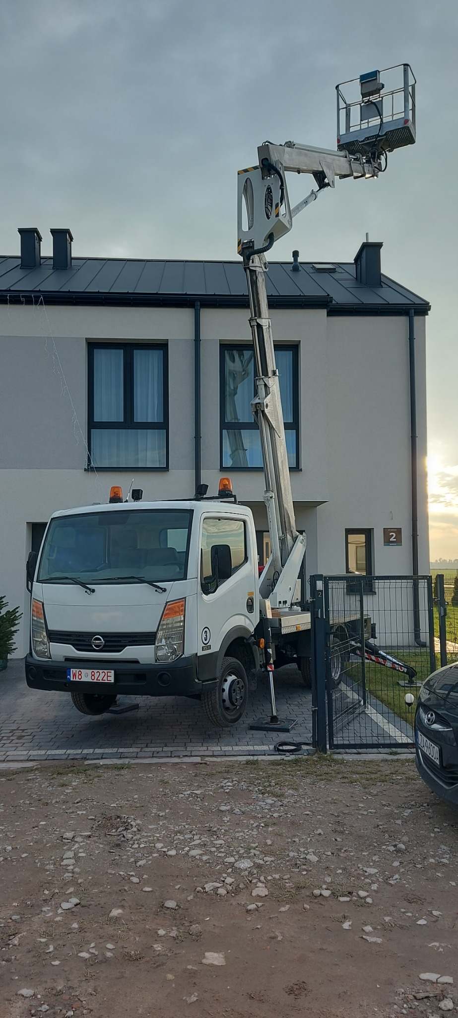 Podnośnik koszowy na tle dwukondygnacyjnego domu z szarą elewacją i ciemnymi oknami. Kosz podnośnika ustawiony przy dachu. Widoczne lampki dekoracyjne.