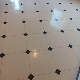 KMV-REMONTY VOLODYMYR KRAVETS - Błyszcząca, kremowa podłoga z kwadratowych płytek z czarnymi wstawkami w romby, ułożona w karo. Widoczne niebieskie taśmy malarskie przy ścianie.