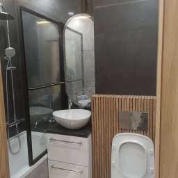 KMV-REMONTY VOLODYMYR KRAVETS - Nowoczesna łazienka z czarnymi płytkami, kabiną prysznicową typu walk-in, umywalką nablatową i drewnianą zabudową toalety. Lustro zabezpieczone folią ochronną.