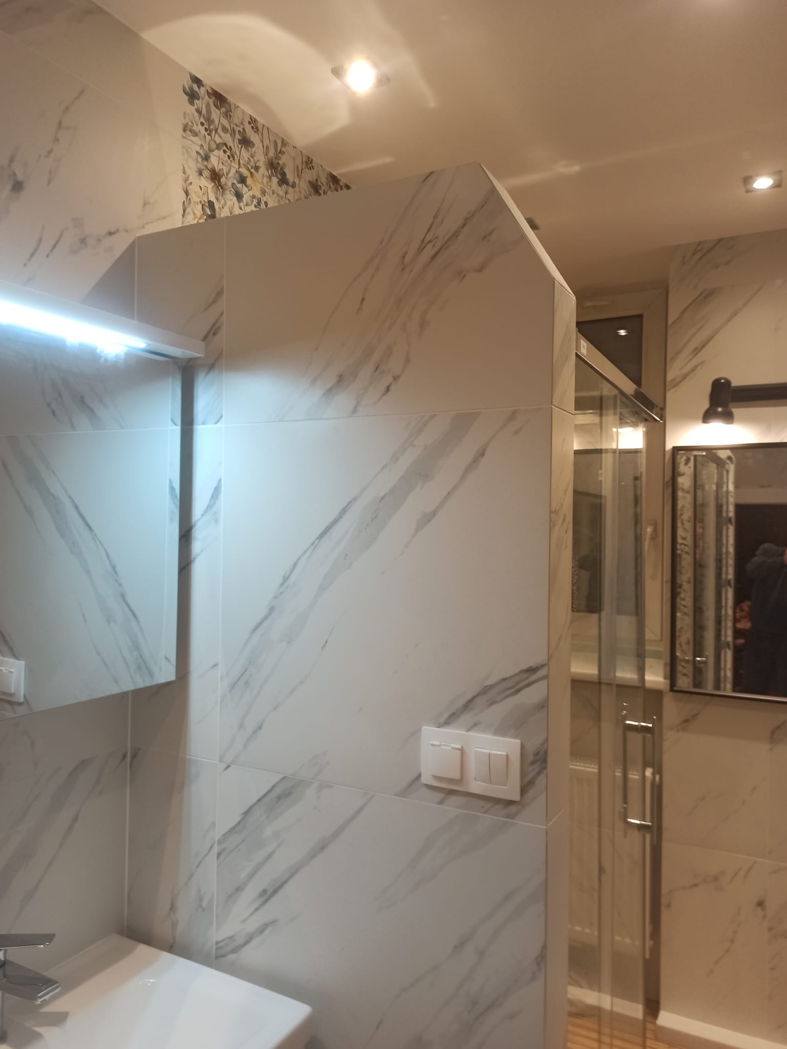 Wykończona łazienka z kabiną prysznicową i umywalką. Ściany wyłożone płytkami imitującymi marmur, akcent kwiatowy na górze. Nowoczesne oświetlenie i armatura.