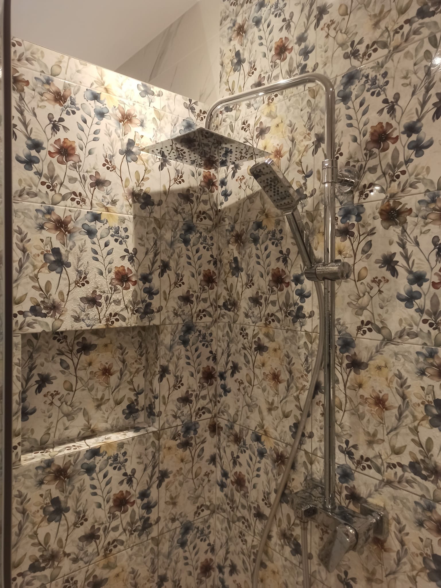 Kabina prysznicowa z deszczownicą i słuchawką, wykończona płytkami w kwiatowy wzór. W tle widoczna półka wnękowa, również wyłożona płytkami.