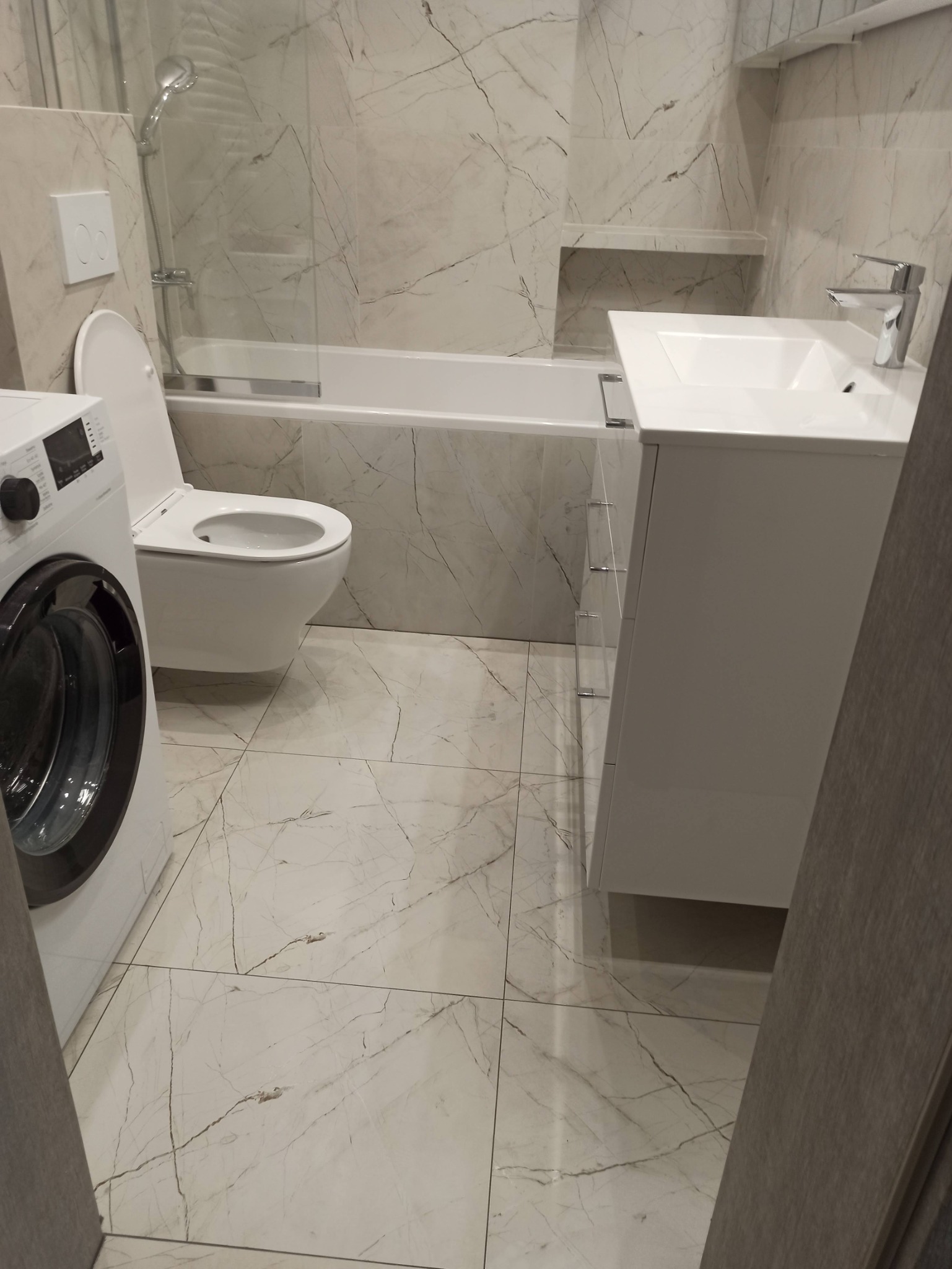 Nowoczesna łazienka z marmurowymi płytkami, białą armaturą i pralką. Umywalka z szafką, toaleta i wanna za szklanym parawanem. Minimalistyczny design.