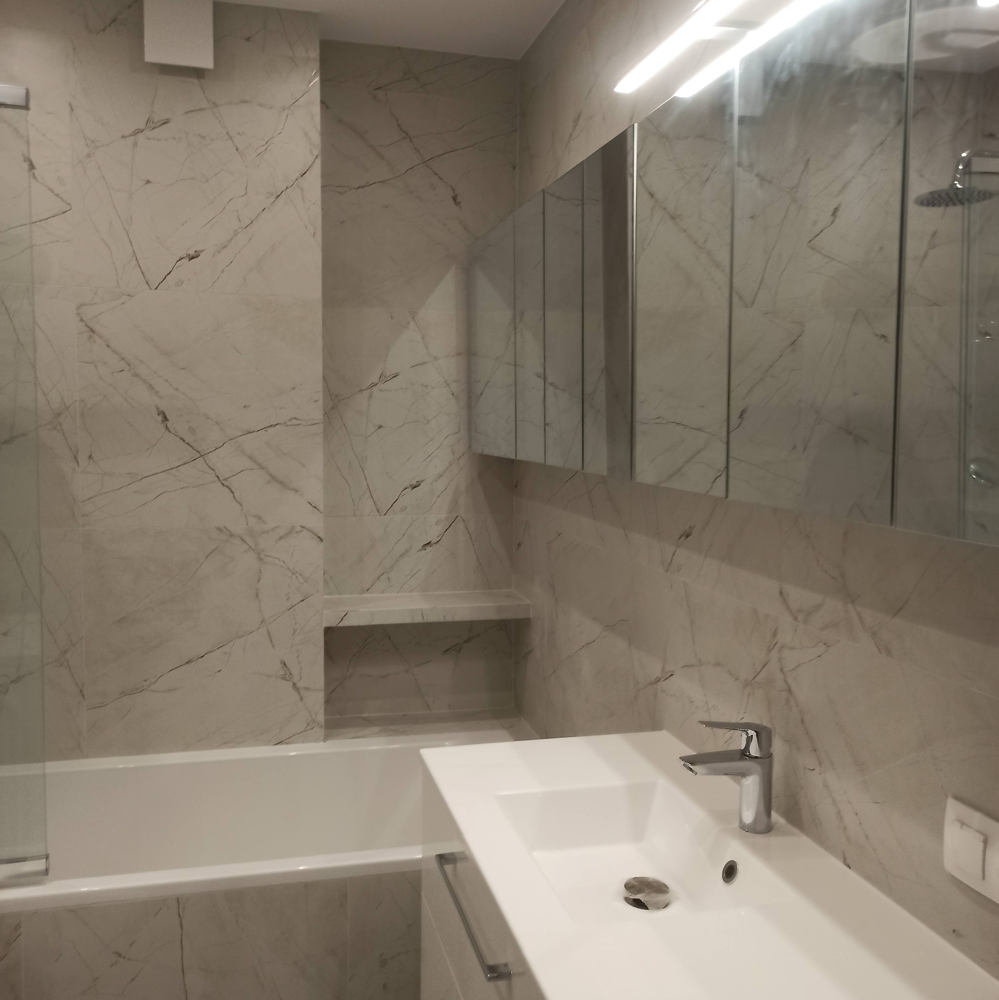 Wykończona łazienka z wanną, umywalką i lustrem w szafce. Ściany i obudowa wanny wyłożone płytkami imitującymi marmur. Minimalistyczny styl, jasne oświetlenie.