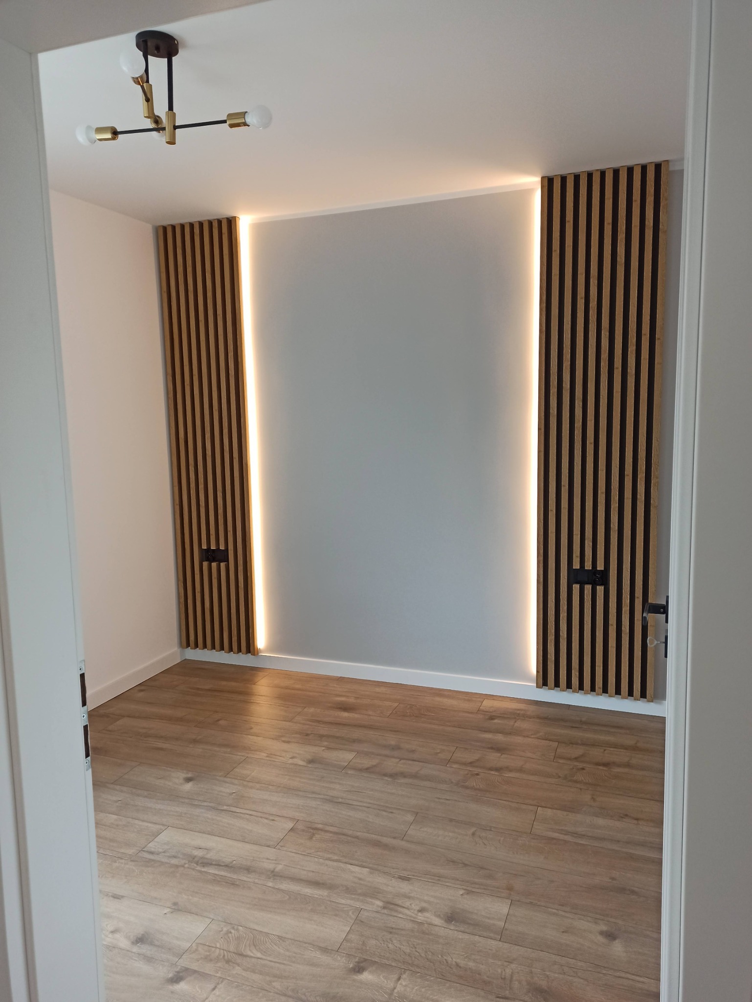 Nowoczesne wykończenie wnętrza: drewniane lamele z podświetleniem LED na ścianie, parkiet na podłodze i minimalistyczna lampa sufitowa.