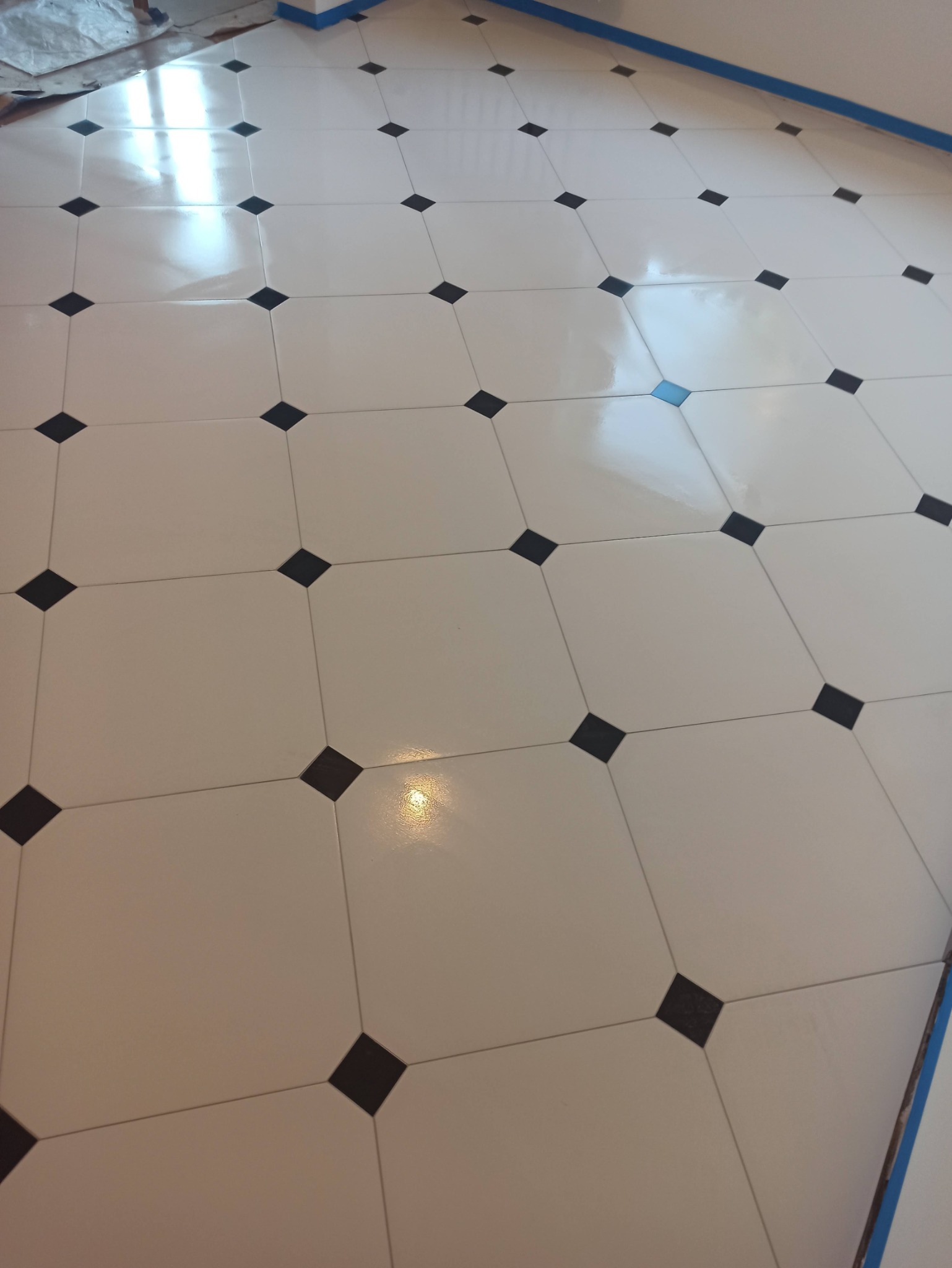 Błyszcząca, kremowa podłoga z kwadratowych płytek z czarnymi wstawkami w romby, ułożona w karo. Widoczne niebieskie taśmy malarskie przy ścianie.