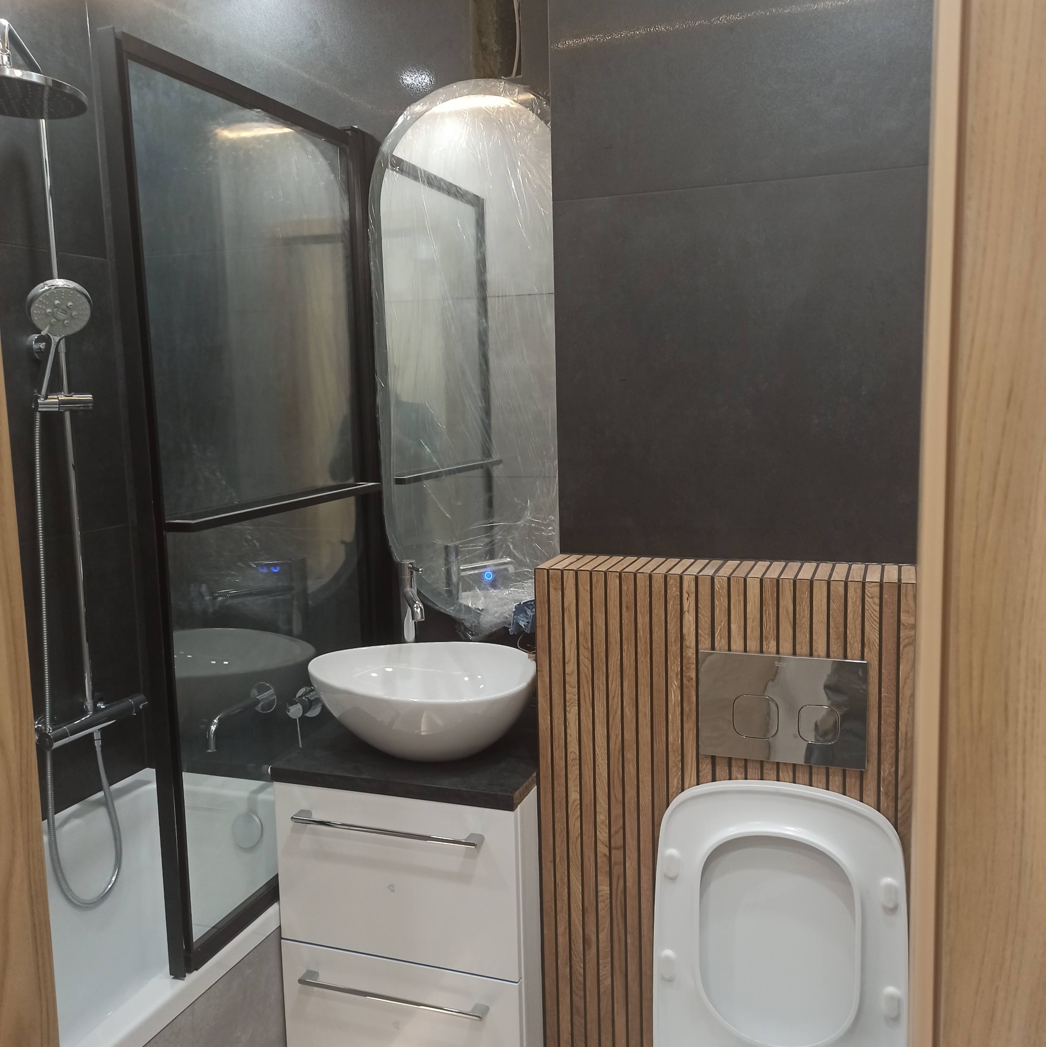 Nowoczesna łazienka z czarnymi płytkami, kabiną prysznicową typu walk-in, umywalką nablatową i drewnianą zabudową toalety. Lustro zabezpieczone folią ochronną.