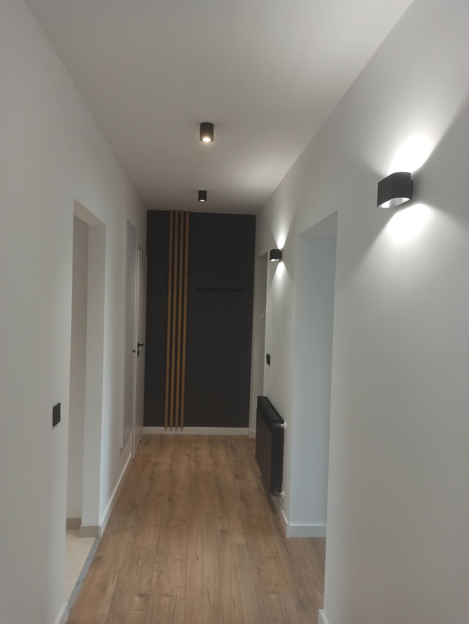 Nowoczesny korytarz z drewnianą podłogą, oświetleniem punktowym i dekoracyjną ścianą w odcieniach szarości z drewnianymi listwami. Minimalistyczny design.