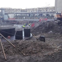 Paweł Stępień - Budowa fundamentów pod konstrukcję przemysłową. Widoczny ciężki sprzęt, zbrojenie, betonowe podpory i pracownicy w odblaskowych kamizelkach. Prace ziemne w toku.