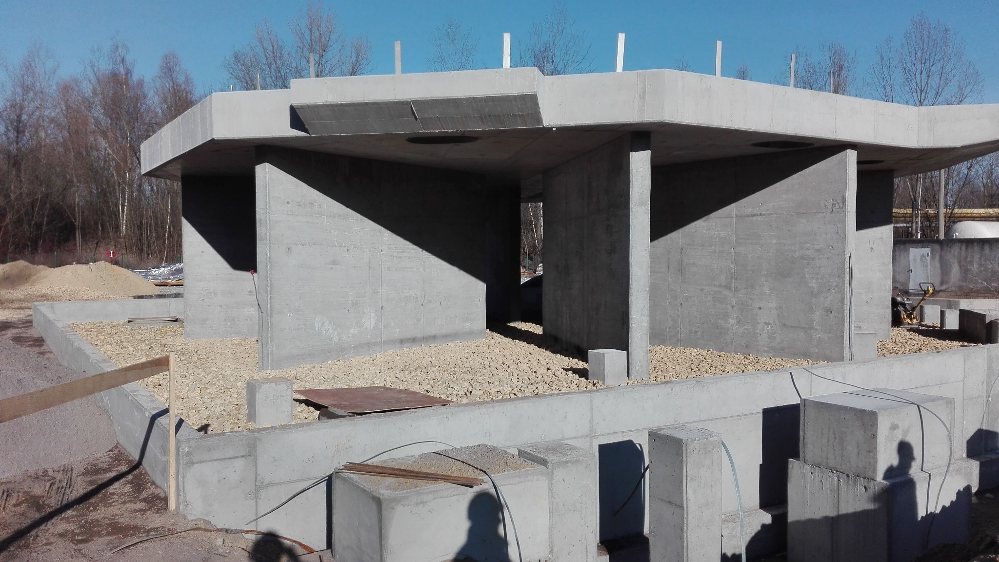 Surowa konstrukcja betonowa w trakcie budowy, widoczne szalunki i fundamenty. W tle drzewa bez liści i błękitne niebo. Elementy zbrojeniowe i podsypka żwirowa.