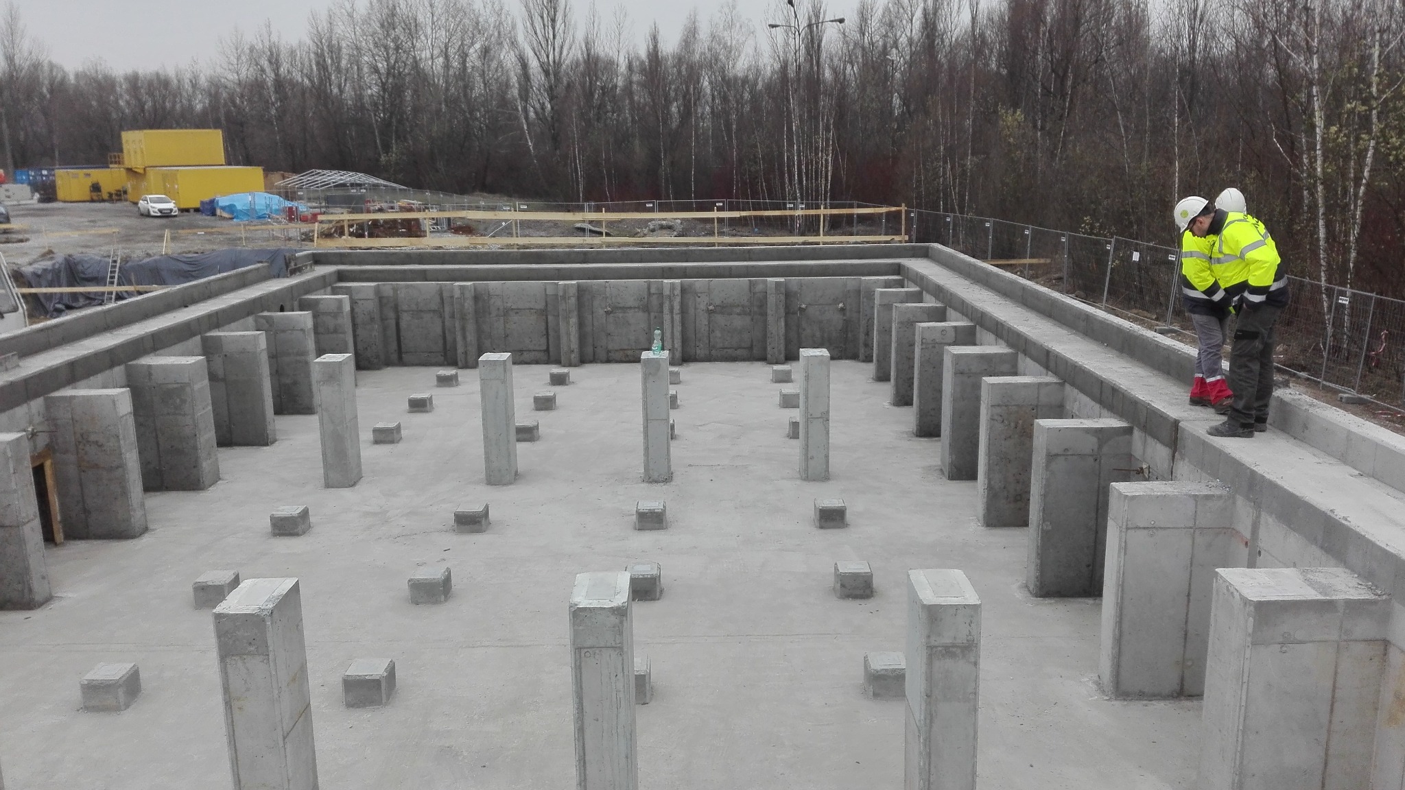 Fundamenty budynku z betonu, widoczne filary i ściany. Dwóch inspektorów w kaskach i jaskrawych kurtkach dokonuje inspekcji. W tle drzewa i kontenery budowlane.