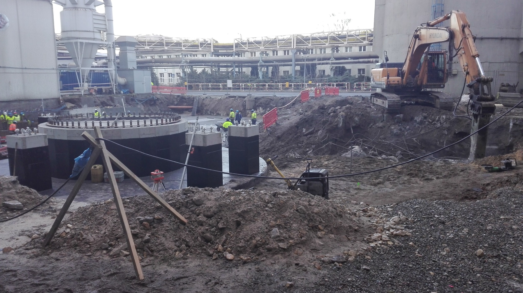 Budowa fundamentów pod konstrukcję przemysłową. Widoczny ciężki sprzęt, zbrojenie, betonowe podpory i pracownicy w odblaskowych kamizelkach. Prace ziemne w toku.