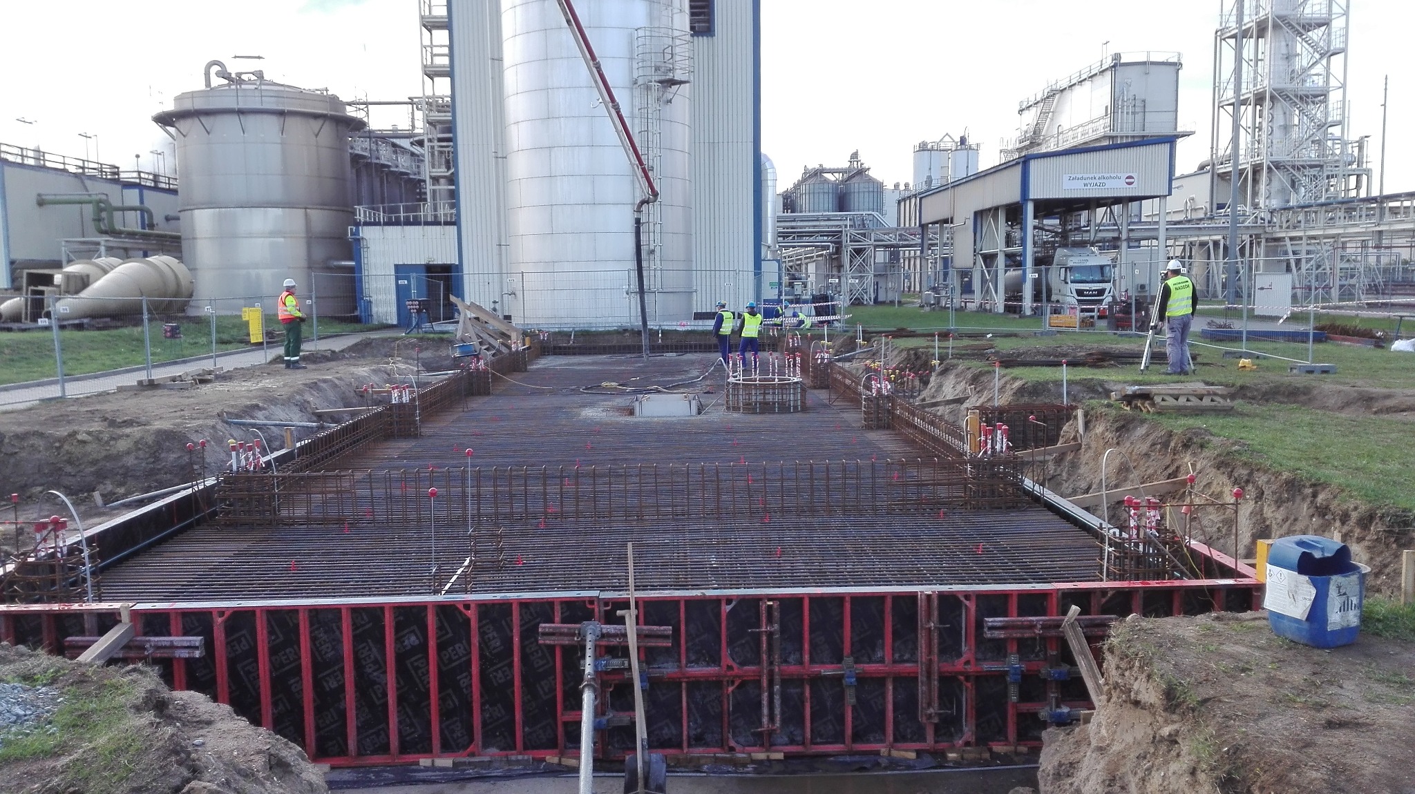 Zbrojenie fundamentu pod budowę przemysłową z widocznymi szalunkami, stalowymi prętami i pracownikami w tle, w trakcie wylewania betonu.