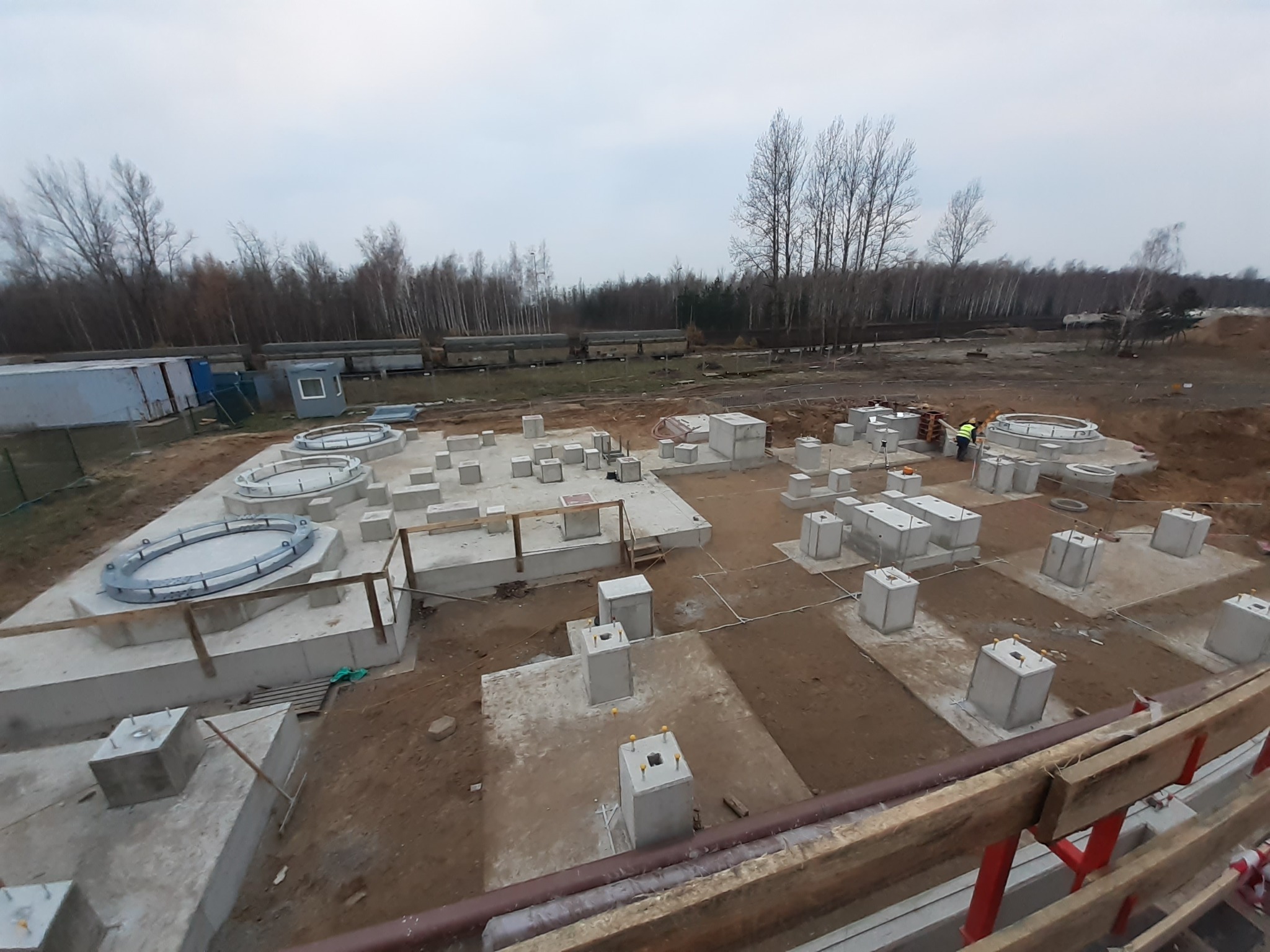 Fundamenty pod budowę z widocznymi betonowymi słupami i pierścieniami. W tle las i pochmurne niebo. Prace budowlane w toku, widoczny pracownik w odblaskowej kamizelce.