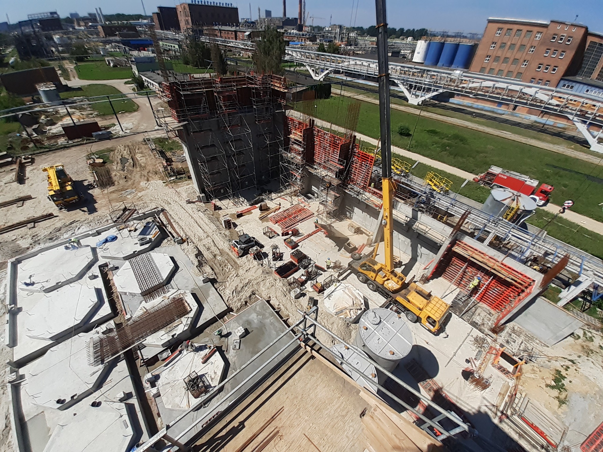 Widok z góry na plac budowy z żurawiem, szalunkami i zbrojeniami. W tle budynki przemysłowe i zieleń. Etap wznoszenia konstrukcji żelbetowej.