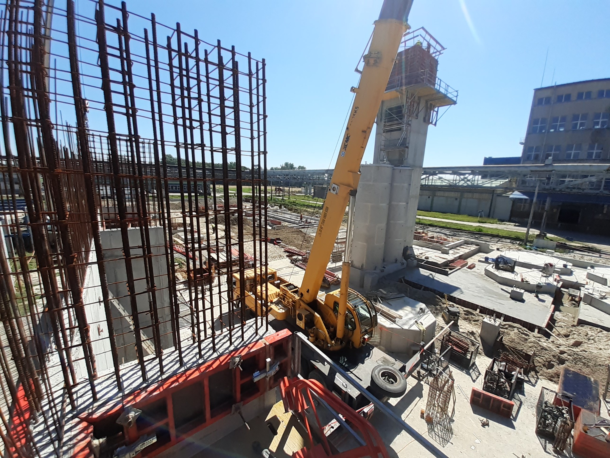 Żuraw na placu budowy z widoczną konstrukcją zbrojeniową i elementami szalunków w słoneczny dzień. Ujęcie z góry, ukazujące postęp prac budowlanych.