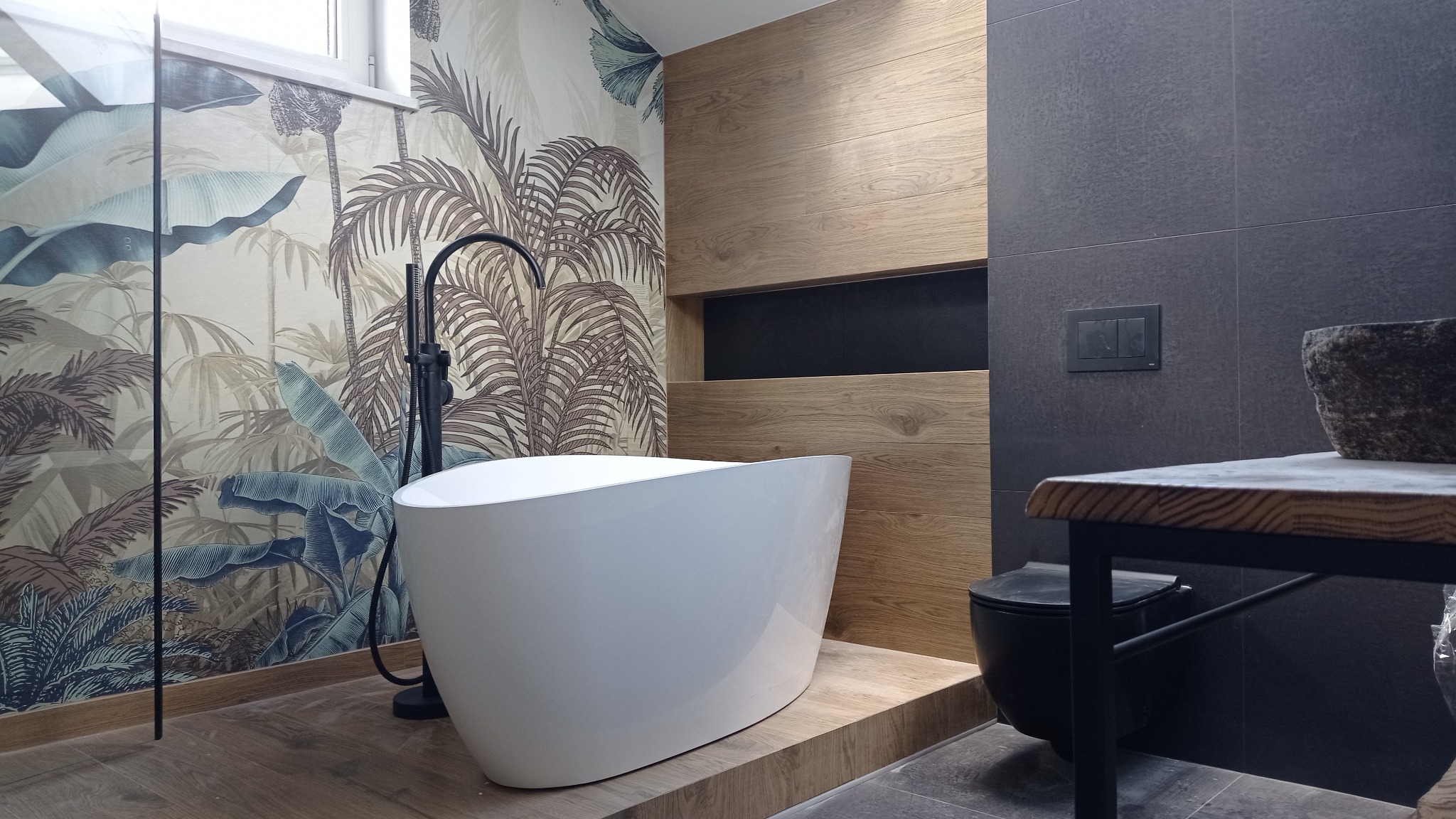 Nowoczesna łazienka z wolnostojącą wanną na podeście, egzotyczna tapeta z motywem roślinnym i drewniane panele ścienne. Minimalistyczny design, czarne akcenty.