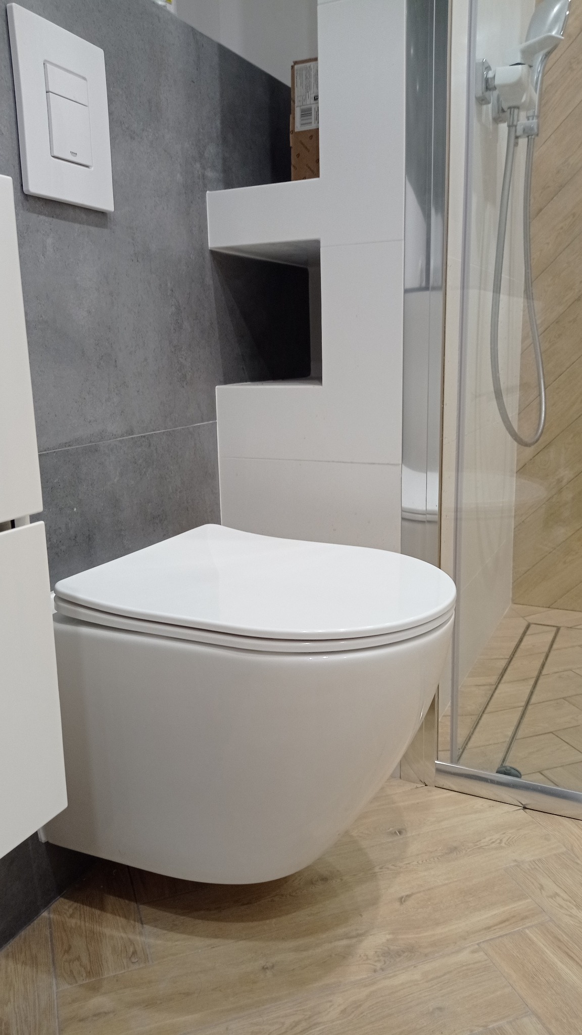 Nowoczesna toaleta podwieszana w wykończonej łazience z szarą ścianą i drewnopodobną podłogą. Widoczna kabina prysznicowa i zabudowa z półkami.