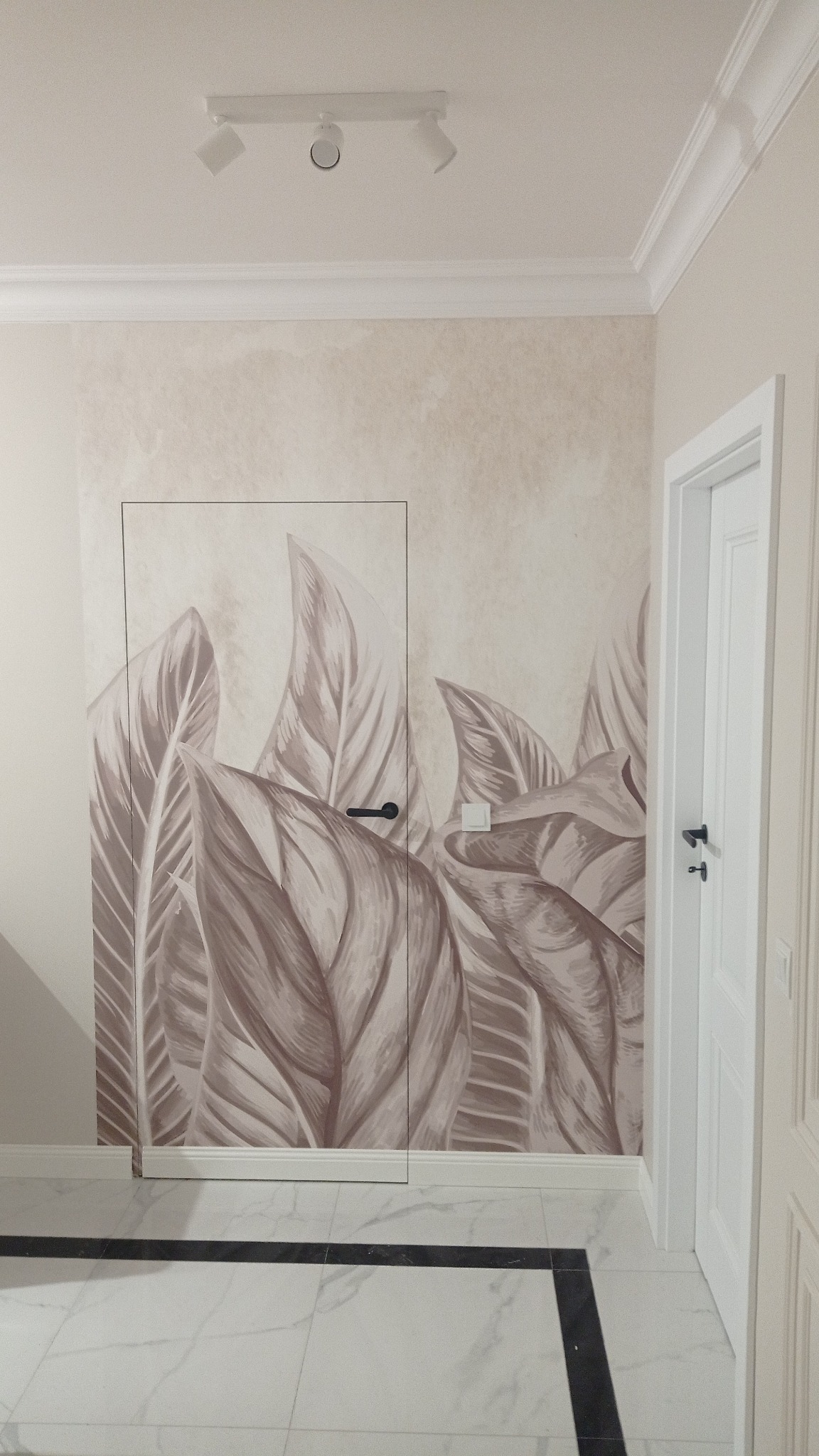Wnętrze z ukrytymi drzwiami w ścianie z motywem liści, białe drzwi po prawej, marmurowa podłoga z czarnym akcentem, oświetlenie sufitowe.