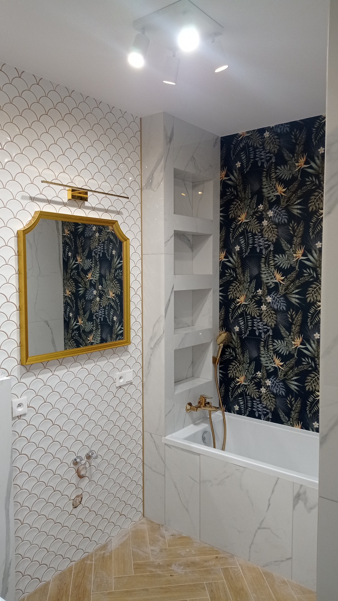 Wykończona łazienka z wanną obudowaną marmurem, półkami w ścianie, lustrem w złotej ramie i wzorzystą tapetą. Na podłodze panele ułożone w jodełkę.