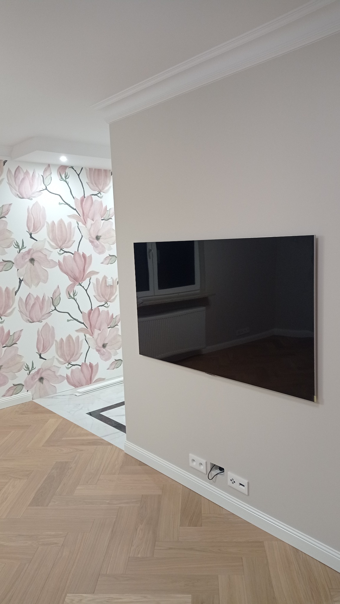 Salon po remoncie: parkiet w jodełkę, ściana z kwiatowym wzorem, minimalistyczny TV na ścianie z ukrytymi kablami i listwy przypodłogowe.