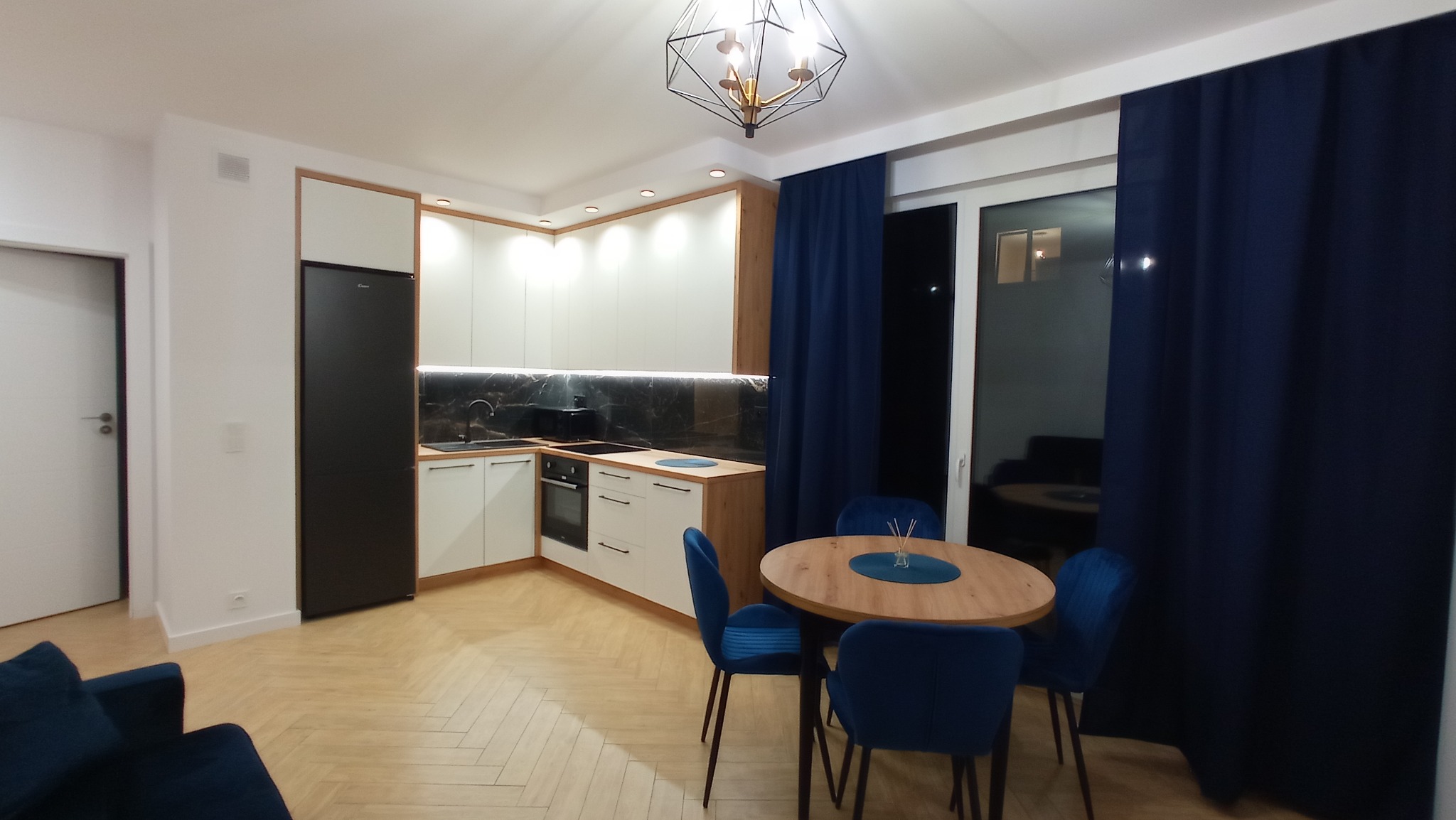 Nowoczesna kuchnia z białymi szafkami, czarnym blatem i lodówką. Parkiet w jodełkę, okrągły stół z niebieskimi krzesłami i ciemne zasłony.