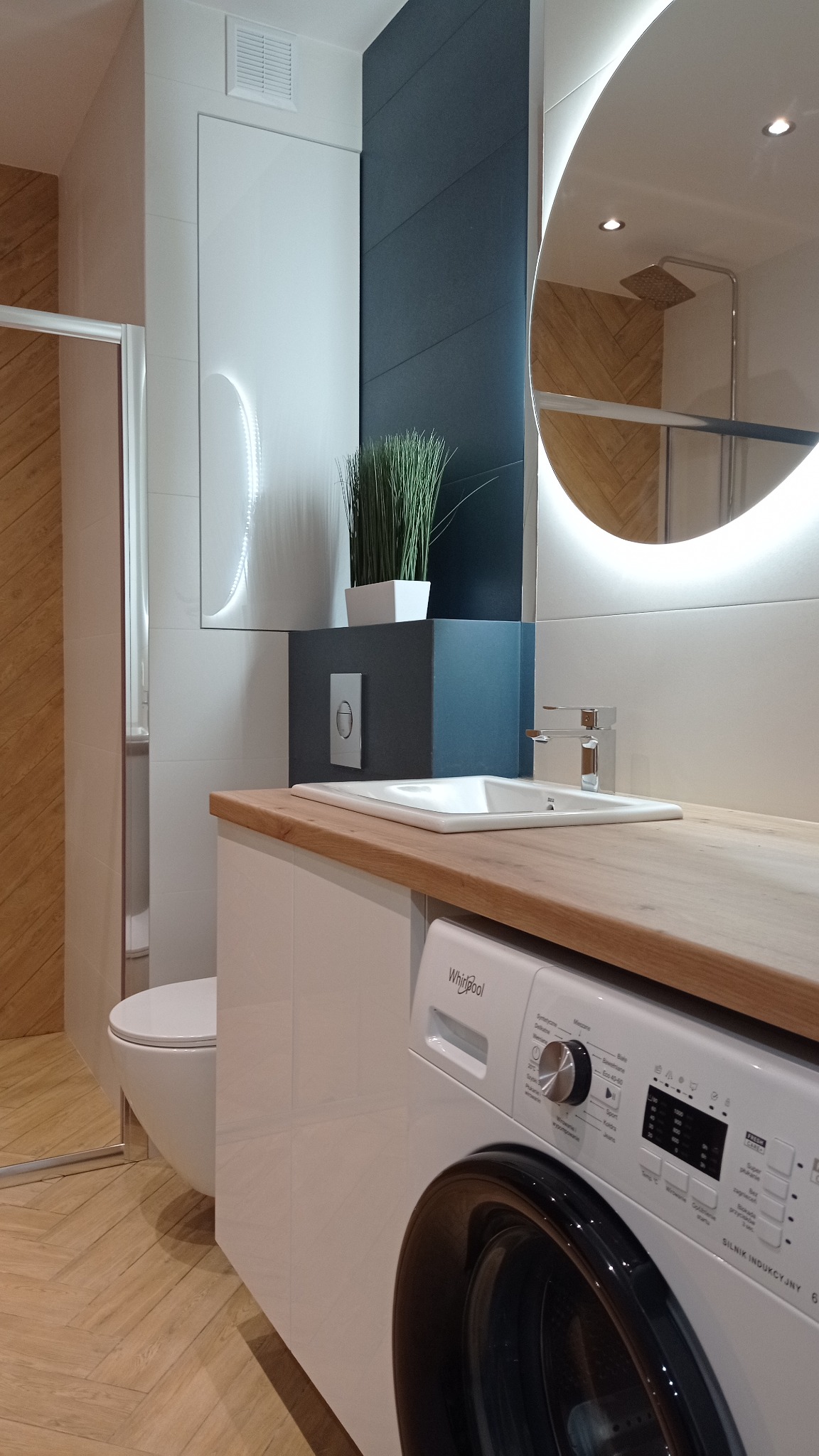 Nowoczesna łazienka z drewnianym blatem, okrągłym lustrem z podświetleniem LED, pralką Whirlpool i płytkami imitującymi drewno na podłodze i w kabinie prysznicowej.