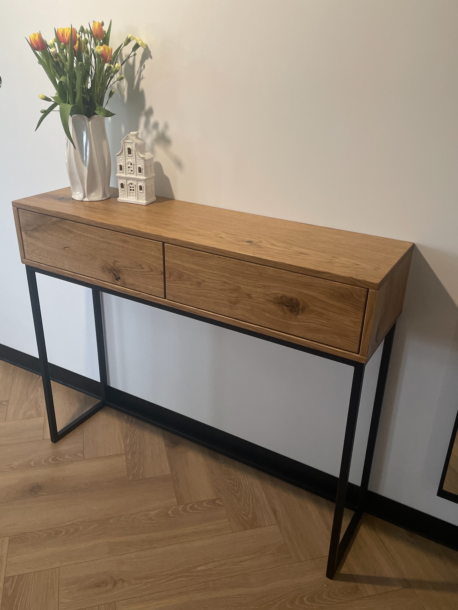 Dębowa konsola z dwiema szufladami na czarnych, metalowych nogach. Na blacie wazon z tulipanami i ceramiczna dekoracja w kształcie domu. Parkiet w jodełkę.