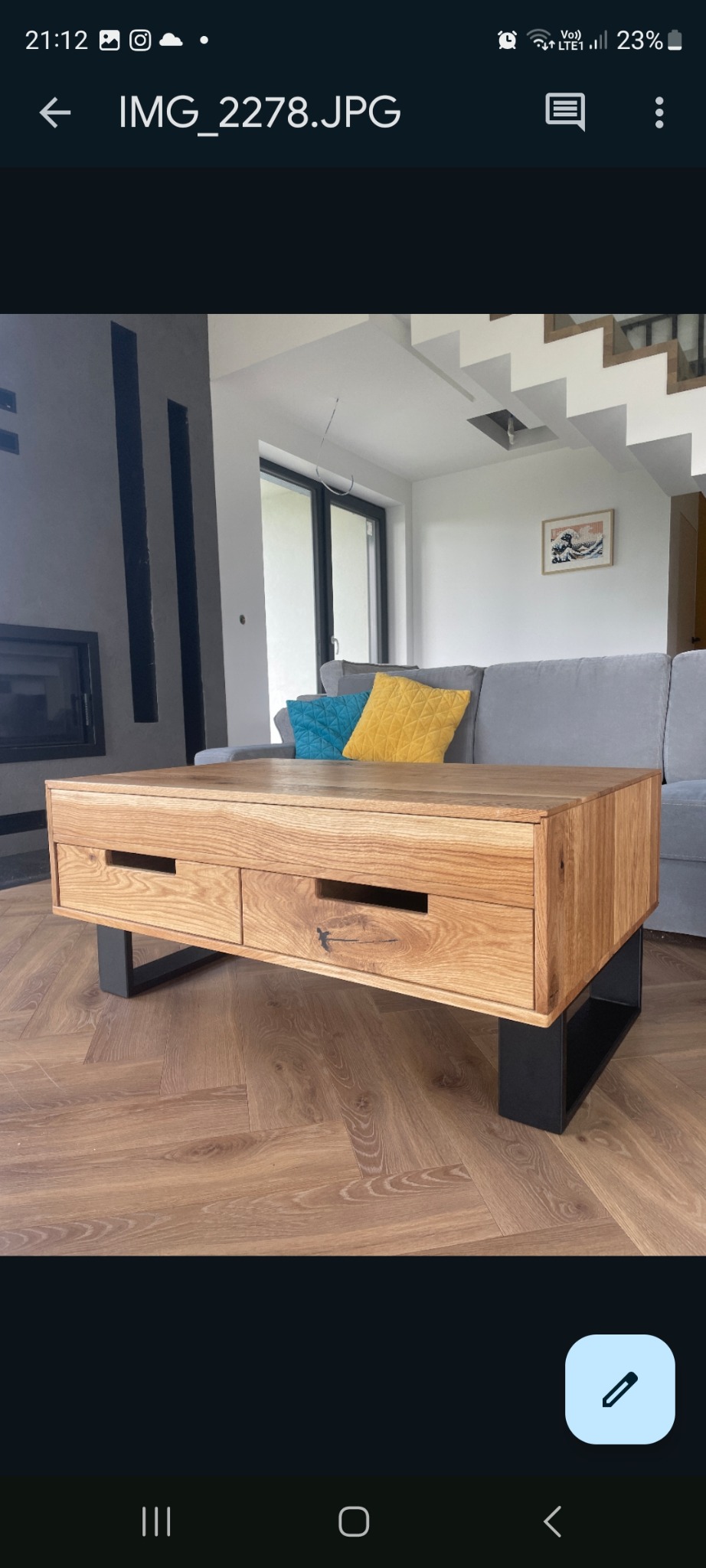 Drewniany stolik kawowy z dwiema szufladami na czarnych, metalowych nogach, na tle salonu z szarą sofą i kominkiem. Parkiet ułożony w jodełkę.