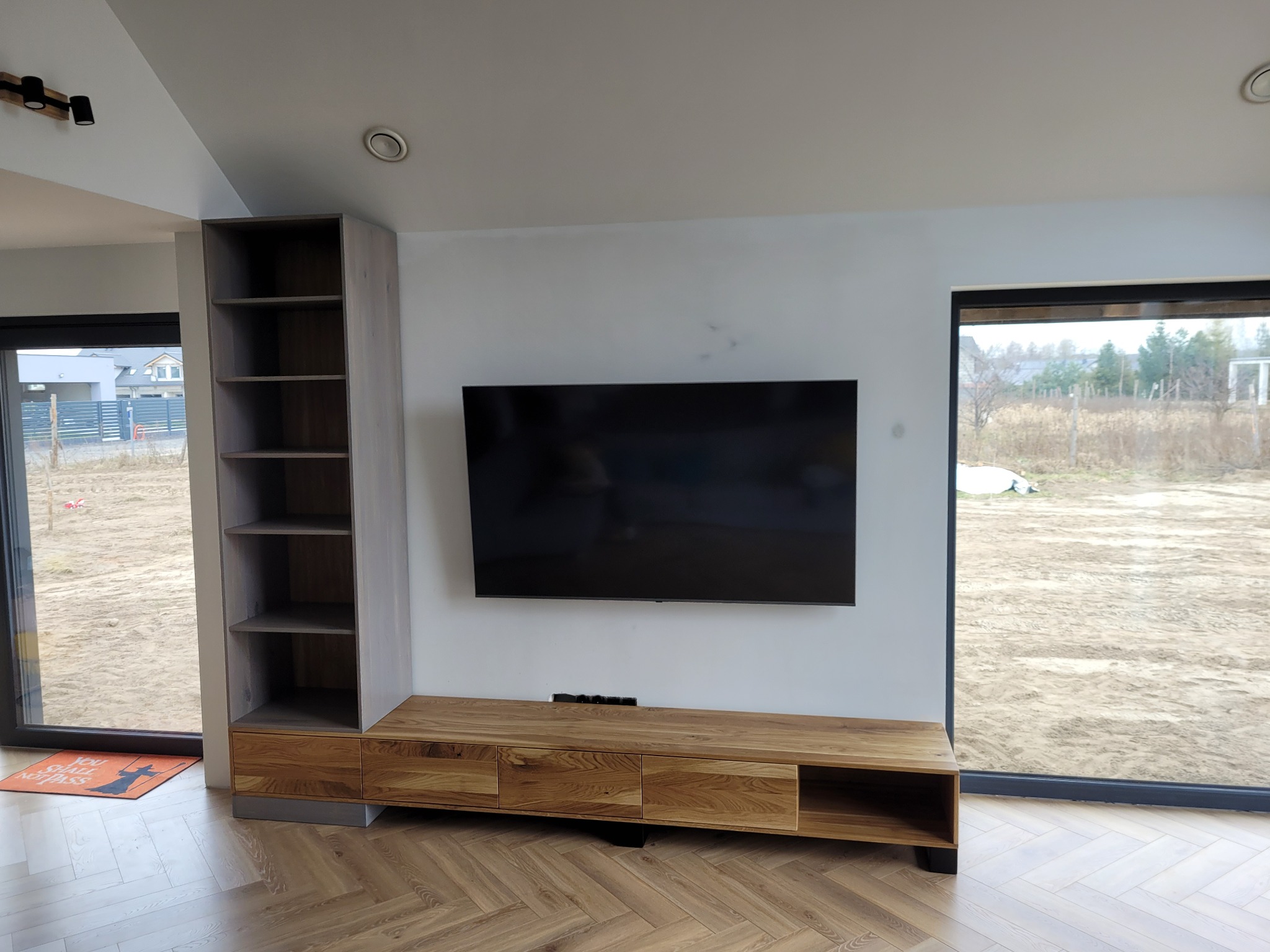 Drewniana szafka RTV z półkami i szufladami, wykonana na zamówienie, z telewizorem na ścianie. Parkiet w jodełkę na podłodze. Widok na zewnątrz przez duże okno.