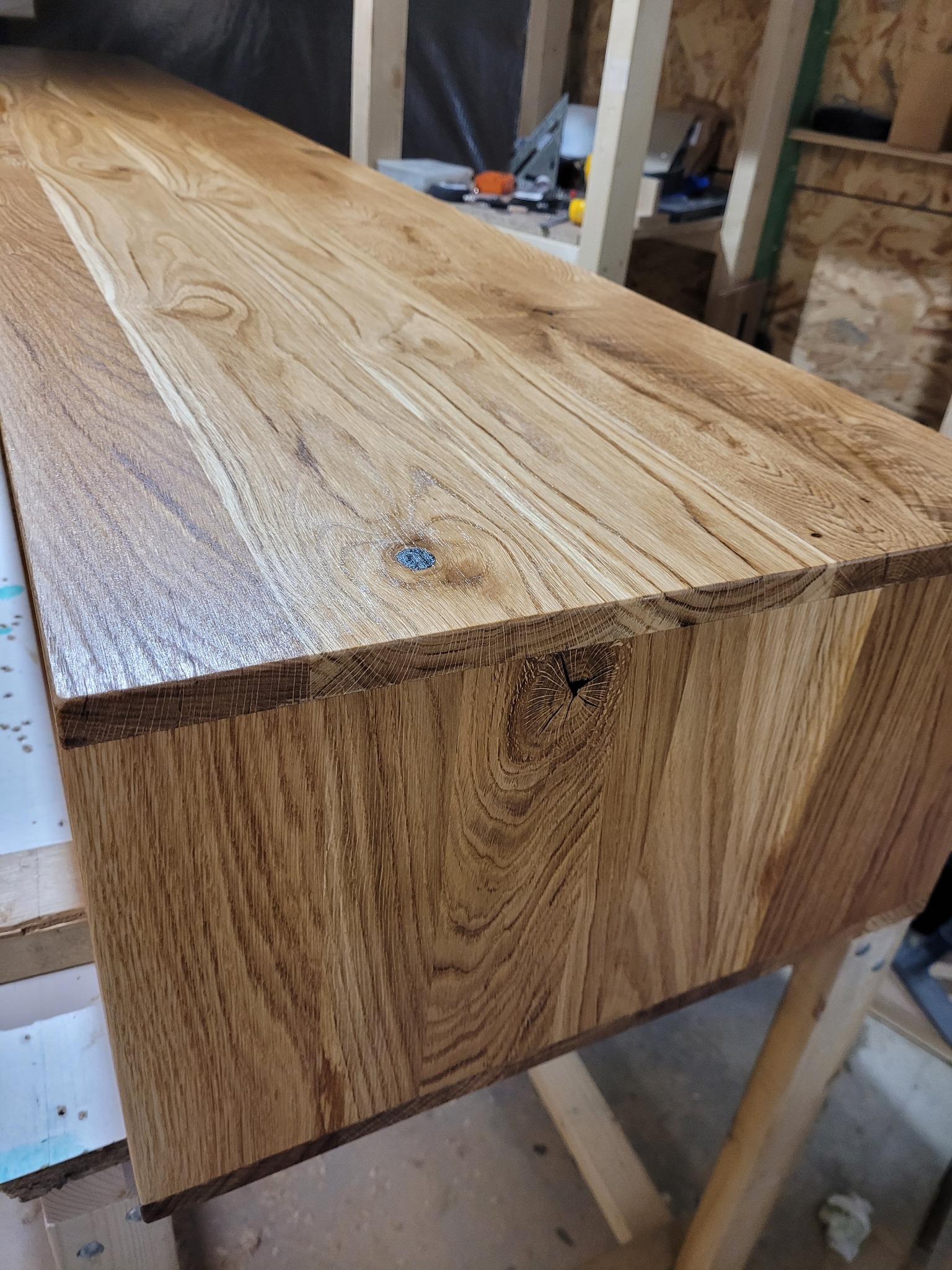 Lakierowany blat dębowy z widocznym usłojeniem i sękiem, na drewnianych kozłach w warsztacie stolarskim. Detal wykończenia krawędzi blatu.