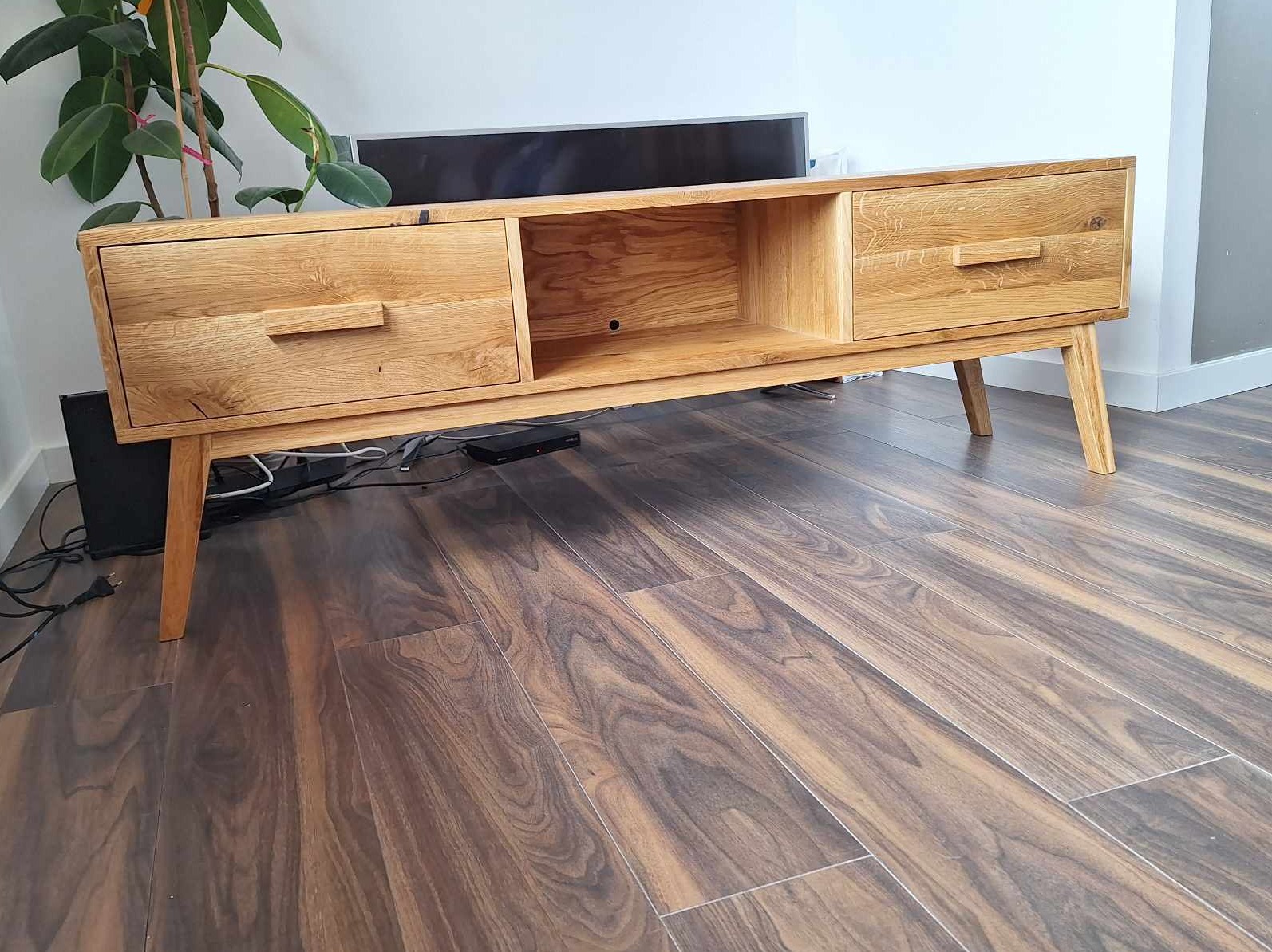 Dębowa szafka RTV w stylu skandynawskim z dwiema szufladami i otwartą półką, ustawiona na drewnianej podłodze. Widoczne kable i sprzęt RTV pod szafką.