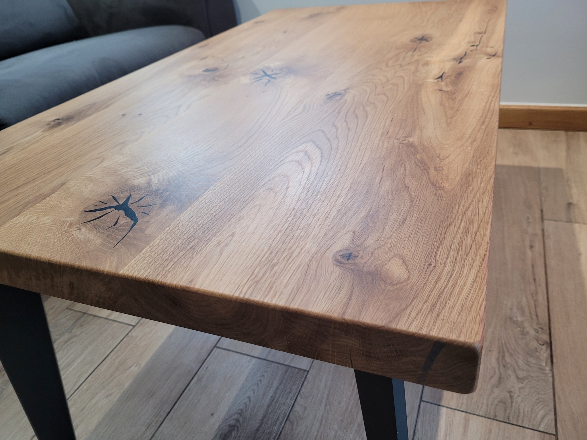 Stół kawowy z blatem z jasnego drewna dębowego, z wyraźnym usłojeniem i czarnymi, metalowymi nogami. Widoczne naturalne sęki i wypełnienia żywicą. Ustawiony na drewnianej podłodze.