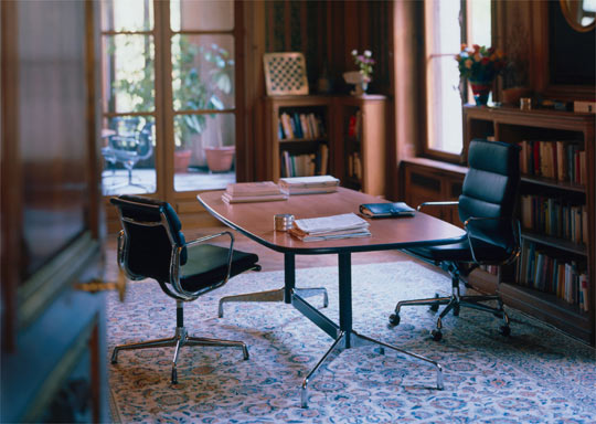 Eames Soft Pad Chairs i stół konferencyjny na tle biblioteczki i okna z widokiem na ogród.