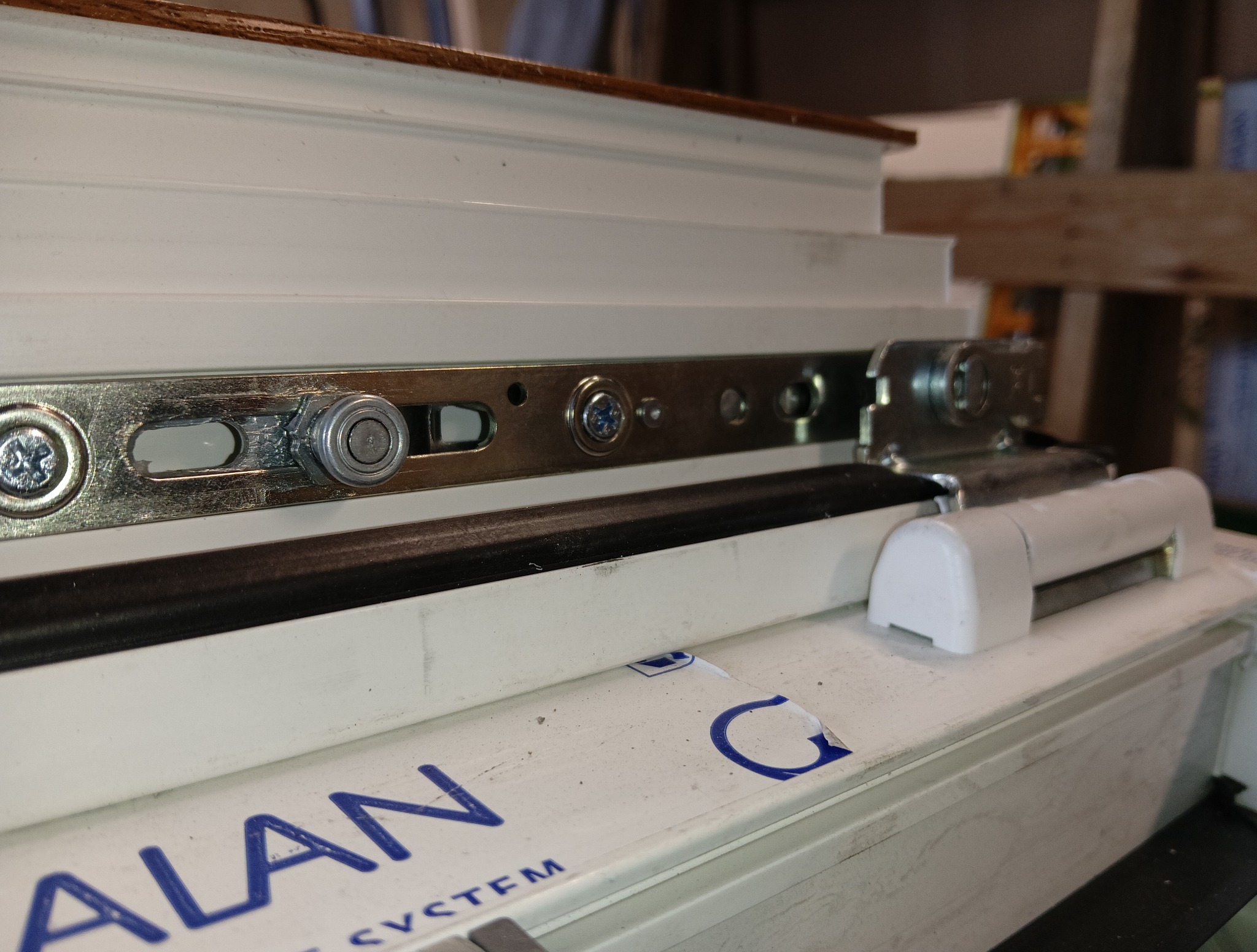 Szczegółowe ujęcie mechanizmu okna PCV marki Alan System, widoczne śruby i metalowe elementy mocujące, zarys drewnianego elementu w tle.