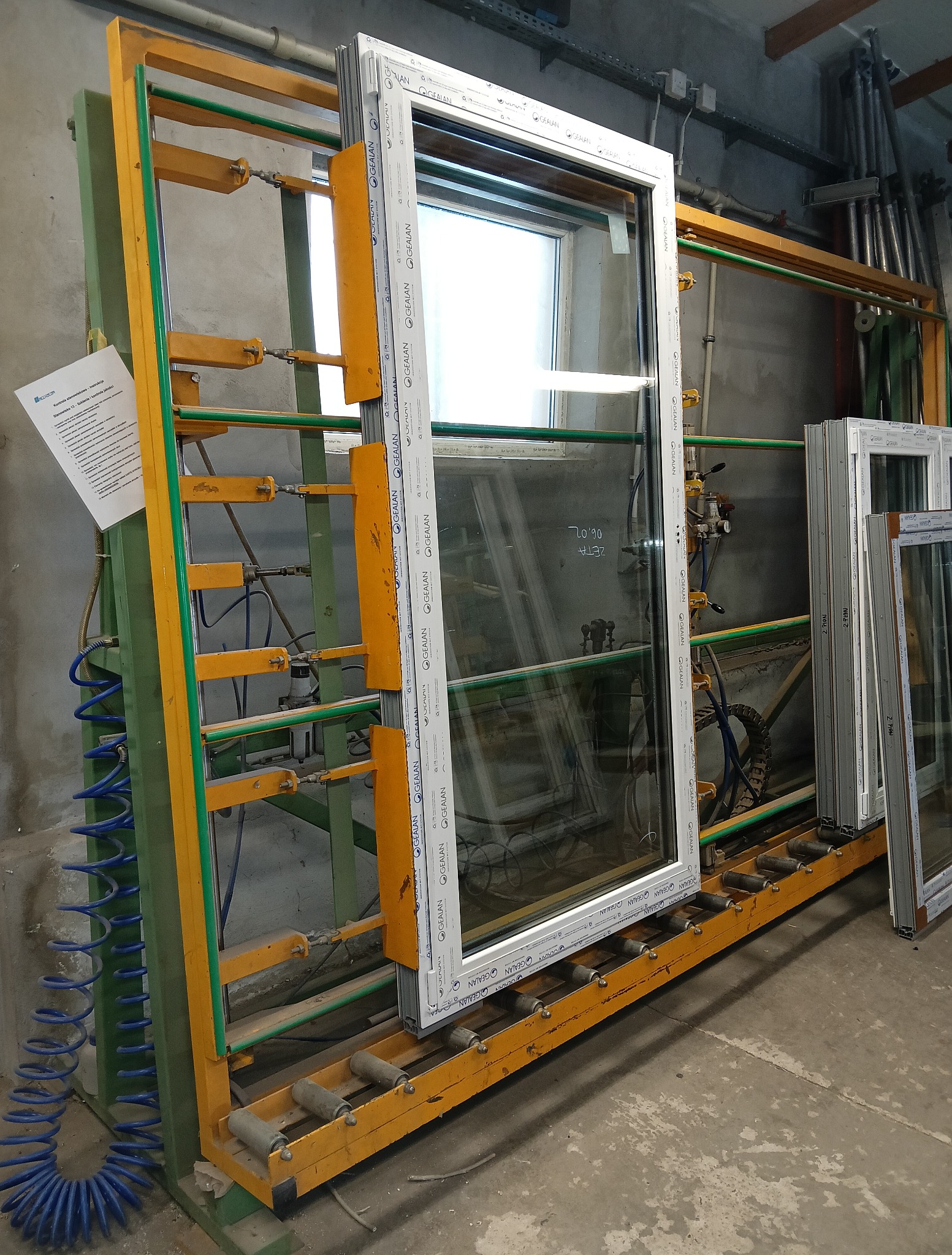 Białe okno PCV marki GEALAN na żółto-zielonym stojaku w hali produkcyjnej. Widoczne inne okna i elementy instalacji. Na oknie notatki 'ZETA 06.02'.