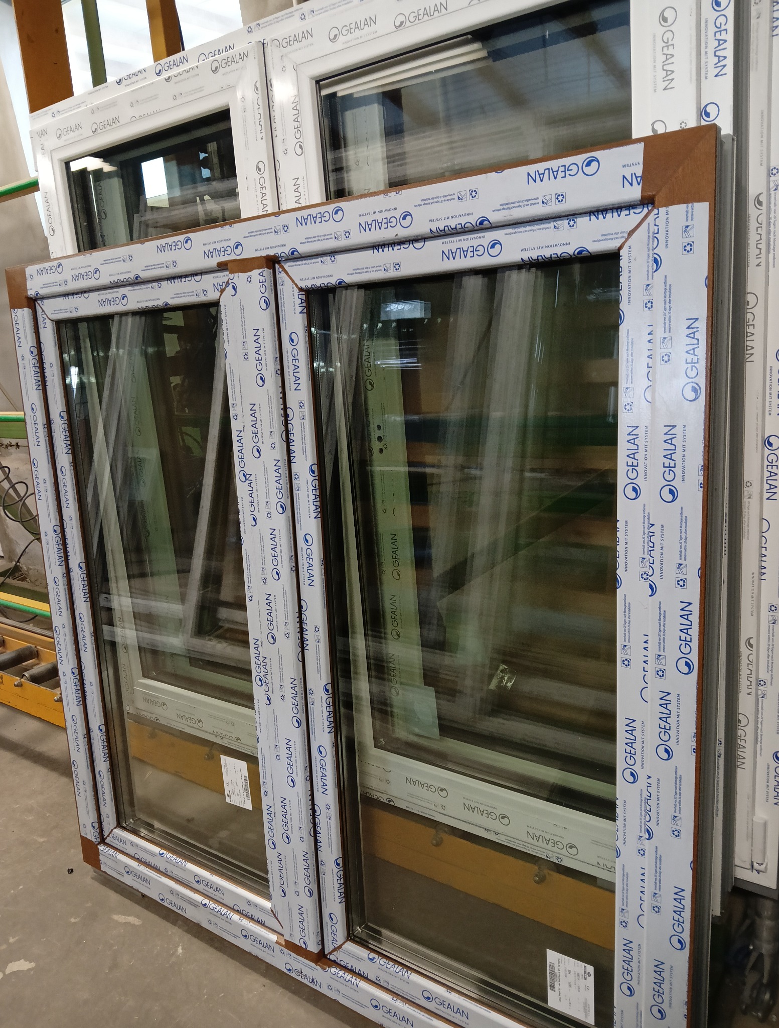 Stos okien PCV Gealan, widoczne profile z folią ochronną. Okna o różnych wykończeniach, w tym imitacja drewna. Widoczne detale konstrukcyjne i etykiety producenta.