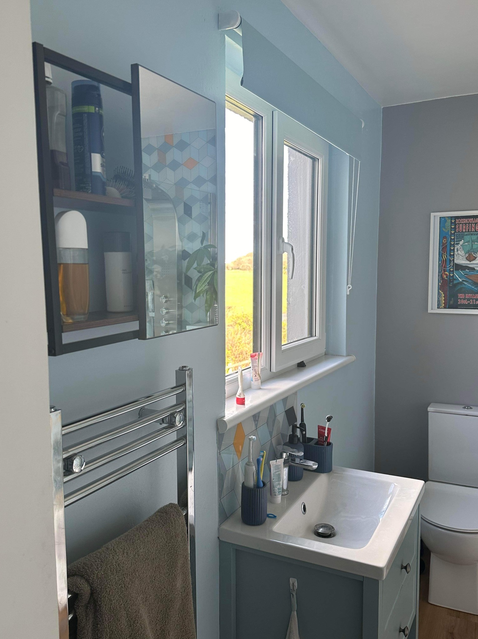 Jasna łazienka z błękitnymi ścianami, umywalką z szafką, grzejnikiem drabinkowym i oknem z widokiem na zieleń. Nad umywalką lustro z geometrycznym wzorem.