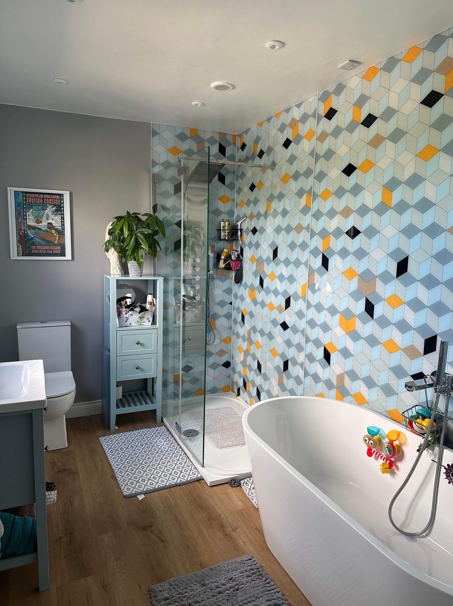 Nowoczesna łazienka z geometrycznymi płytkami w odcieniach szarości, błękitu i żółci. Wolnostojąca wanna, kabina prysznicowa i szafka z rośliną doniczkową.