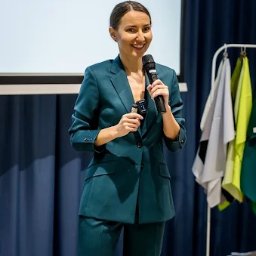 Ilona Sodolska - Ekspert od kolorów w biznesie - Uśmiechnięta kobieta w szmaragdowym garniturze, trzymająca mikrofon, prezentuje na tle ekranu i wieszaka z tkaninami podczas szkolenia.