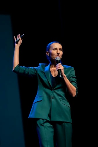 Kobieta w zielonym garniturze przemawia do mikrofonu na scenie, trzymając w ręku pilot prezentera. Ciemne tło, oświetlenie sceniczne.