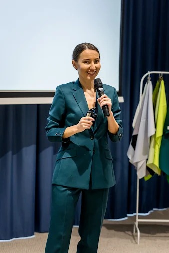 Uśmiechnięta kobieta w szmaragdowym garniturze, trzymająca mikrofon, prezentuje na tle ekranu i wieszaka z tkaninami podczas szkolenia.