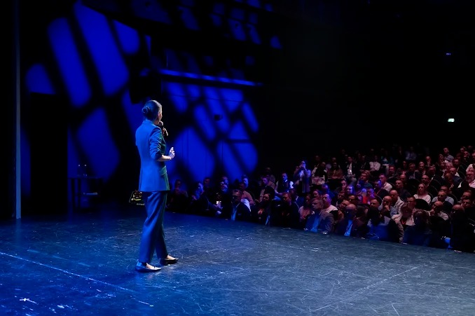 Prelegentka w niebieskim garniturze przemawia do publiczności na scenie oświetlonej niebieskim światłem. Tłum słuchaczy siedzi na widowni.